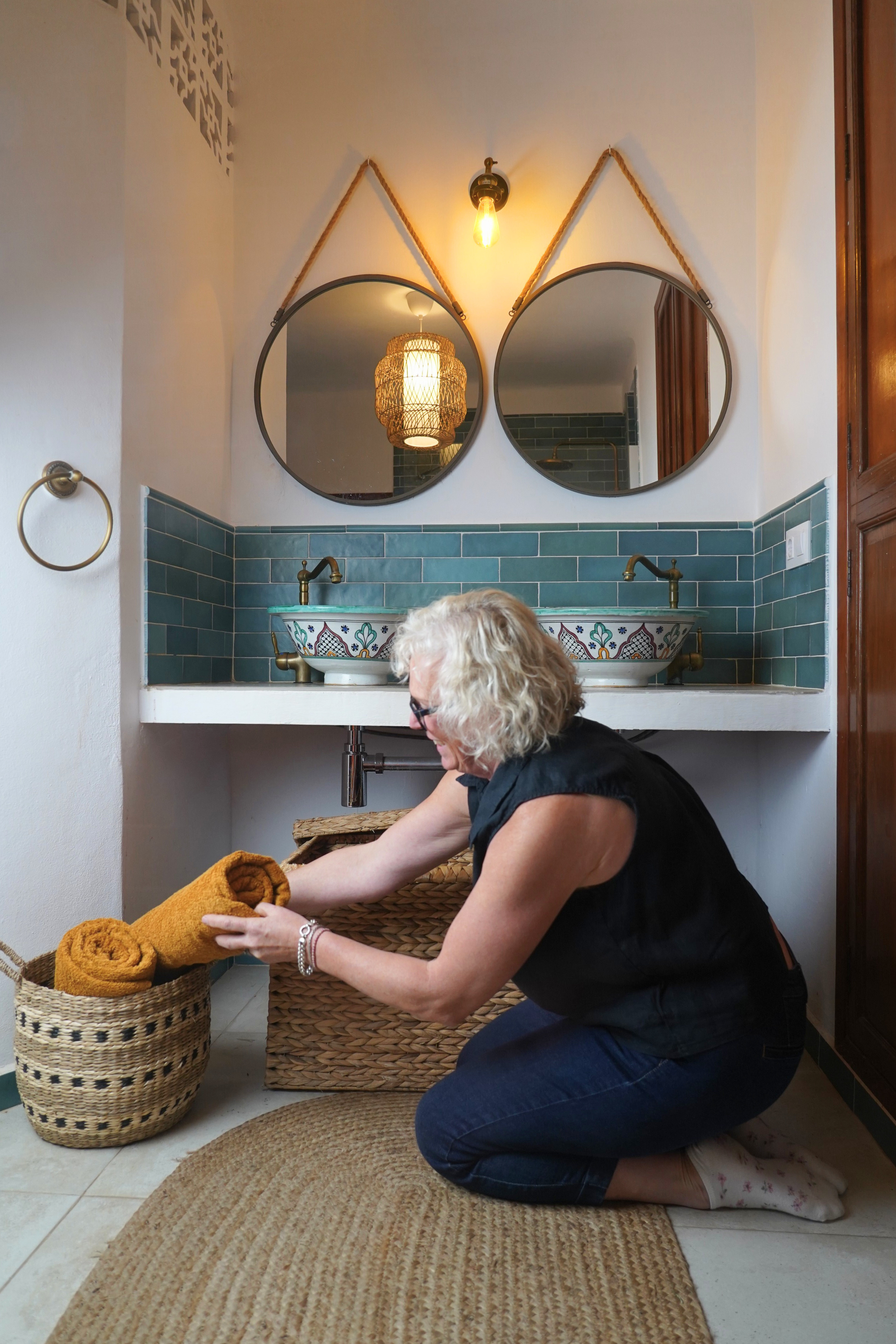 Woman kneeling on a beige rug, folding orange towels from a wicker basket in a bathroom with turquoise tile, white countertop, and unlit wall-mounted mirror.