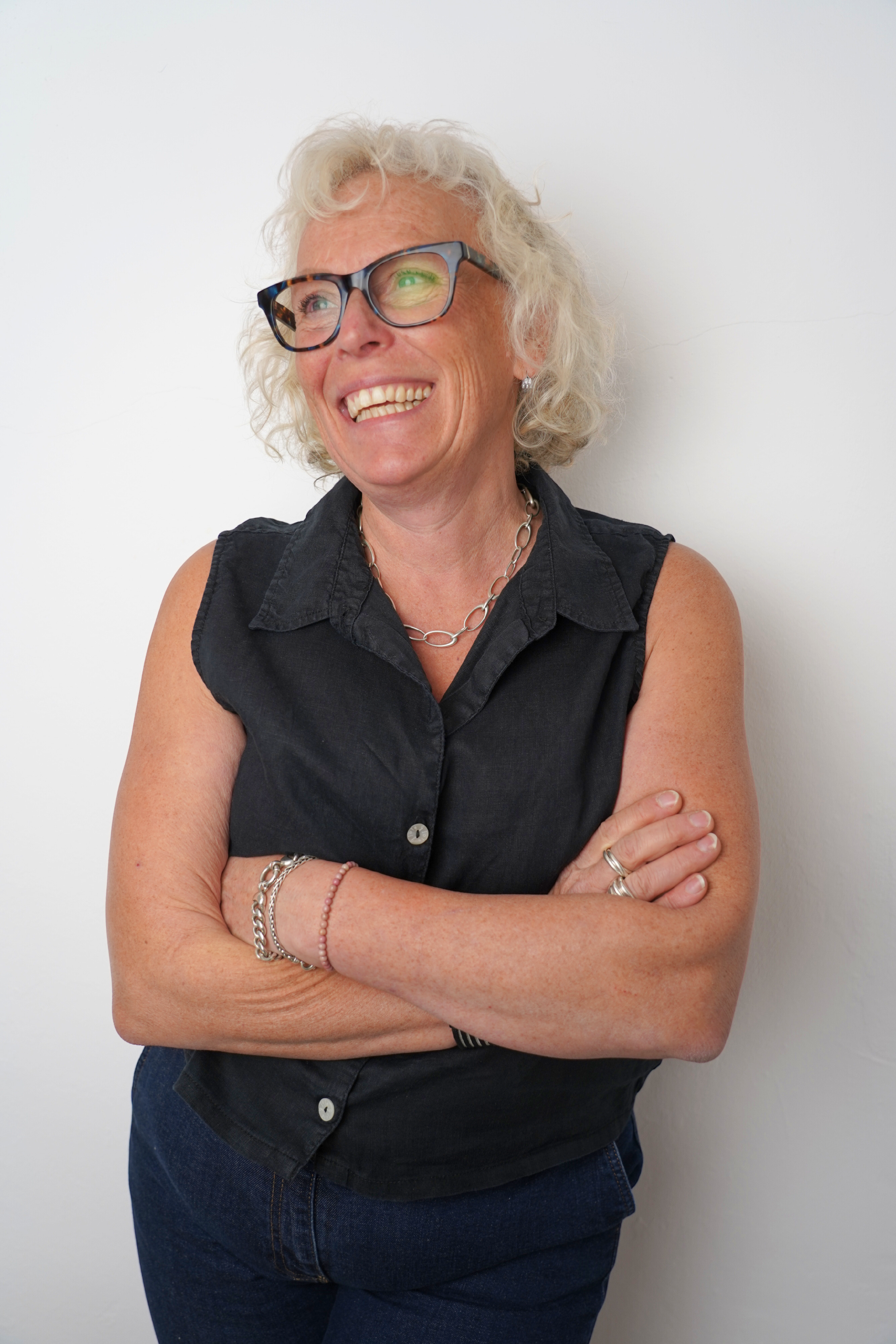 A smiling older woman with curly gray hair, wearing glasses, a sleeveless black shirt, and jewelry, standing against a plain white wall with her arms crossed.