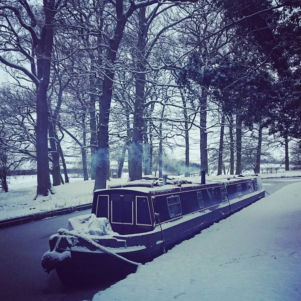 A narrowboat covered in snow docked on a canal with leafless trees in the background.