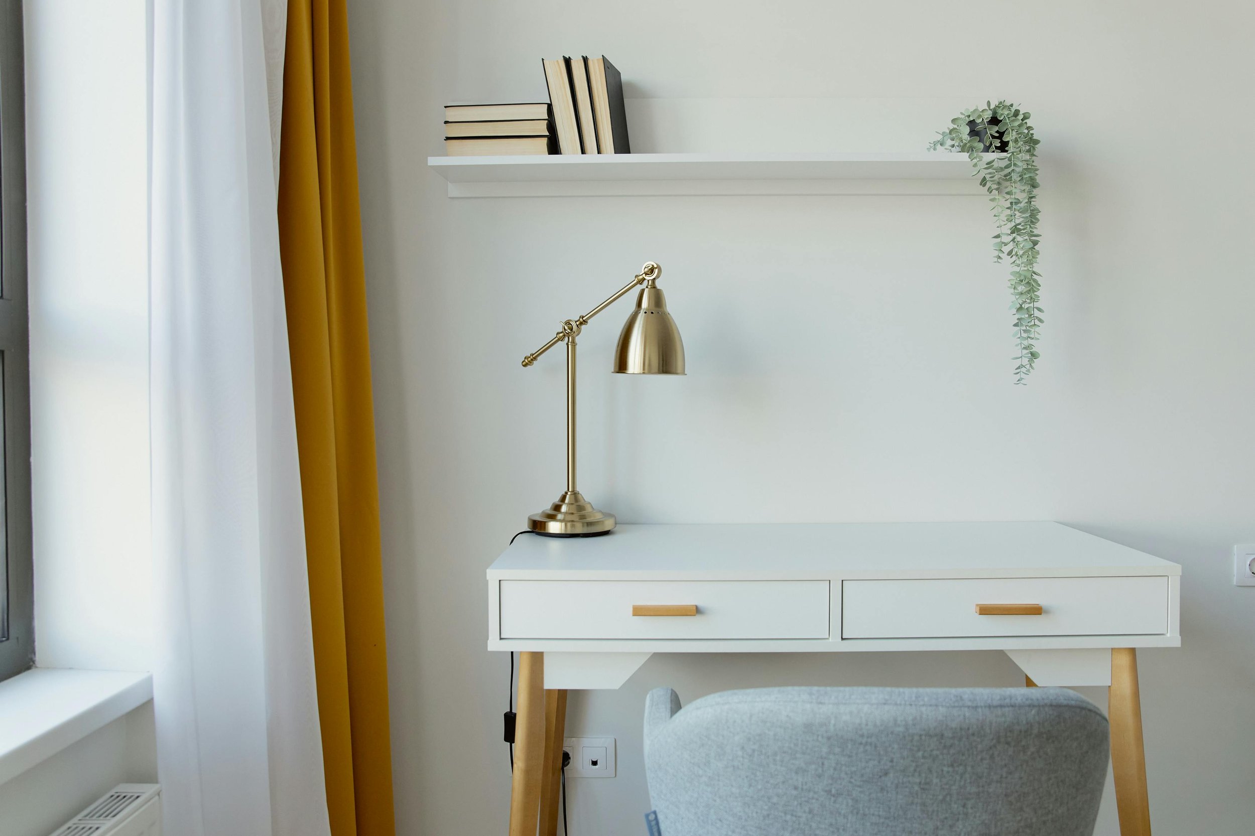 White desk with two drawers and wooden legs, a brass desk lamp, a white wall shelf with books, mustard yellow and white curtains, a gray chair, and a hanging green plant on a white wall.