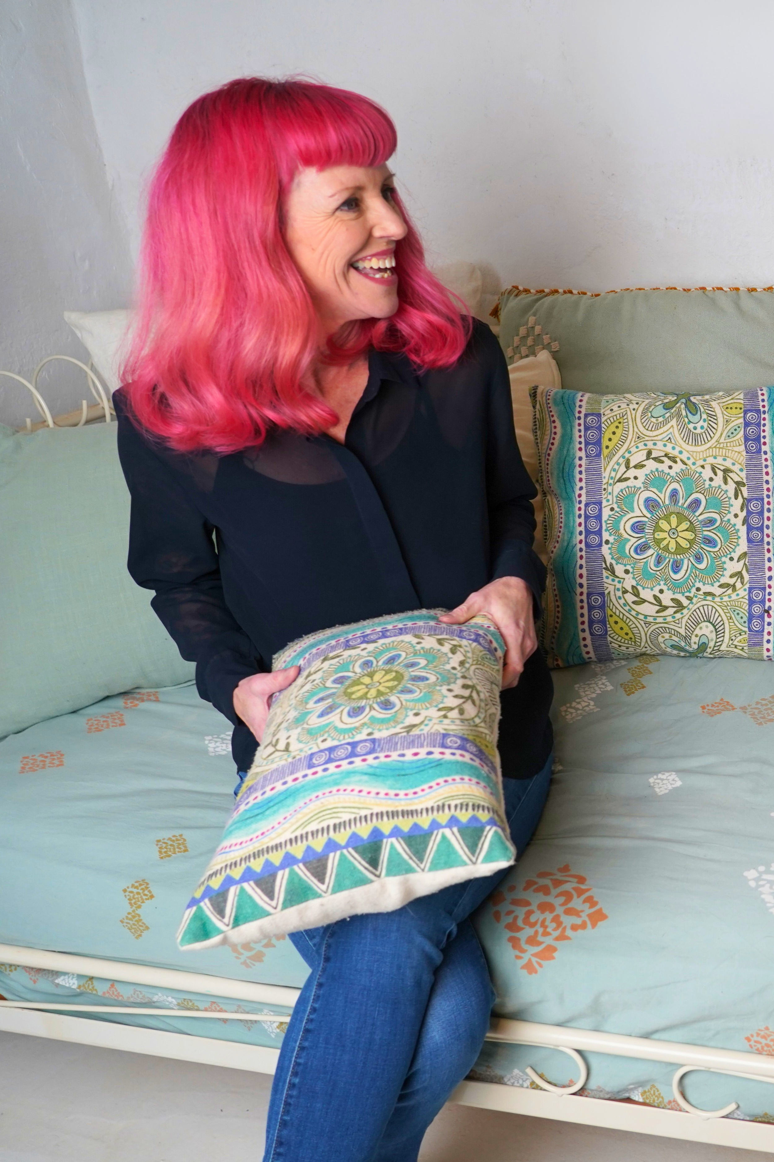 A woman with bright pink, shoulder-length hair, sitting on a bed with decorative pillows, holding a patterned pillow, and smiling while looking to the side.