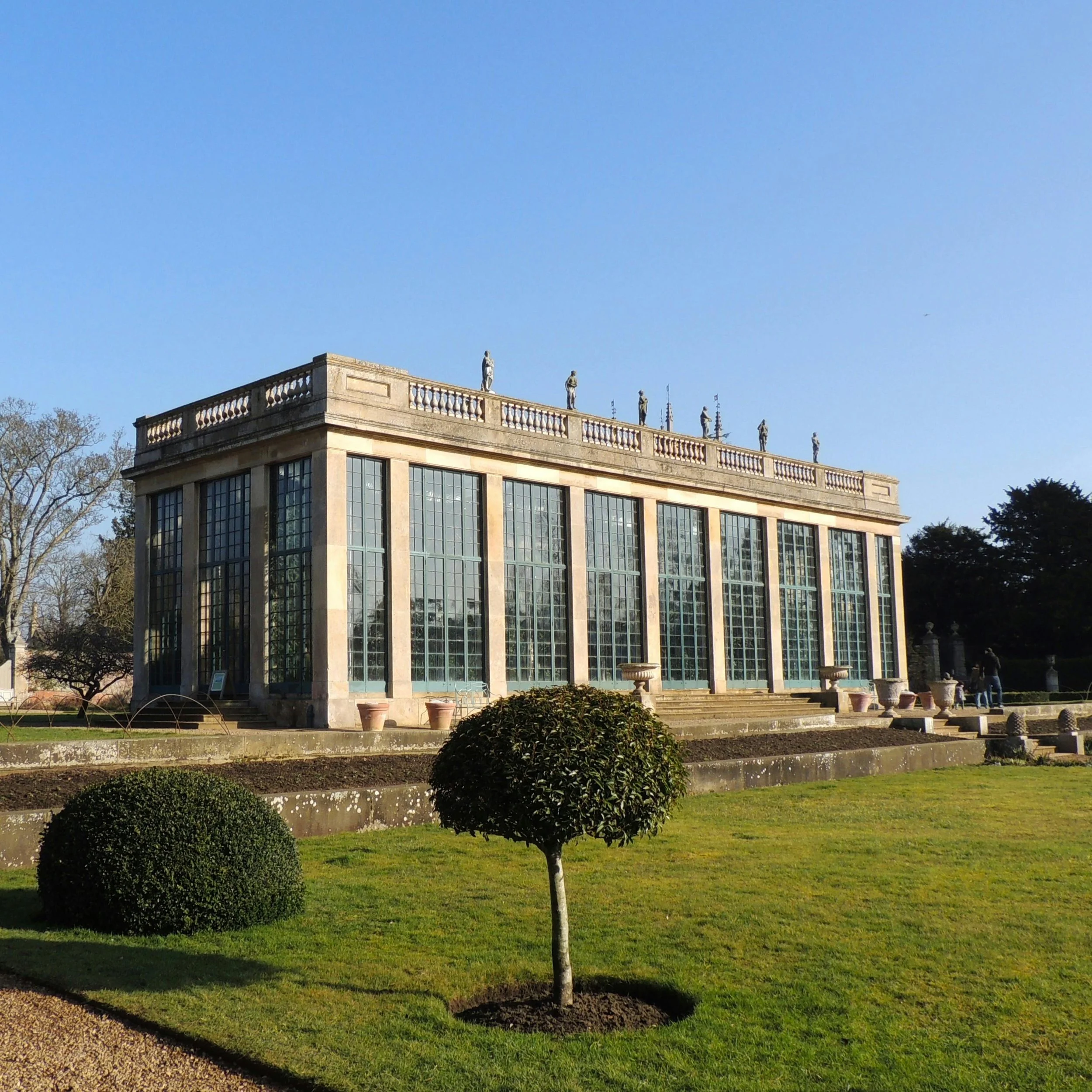 A historic glass and stone building with classical sculptures on the roof, surrounded by manicured lawns and trees under a clear blue sky.