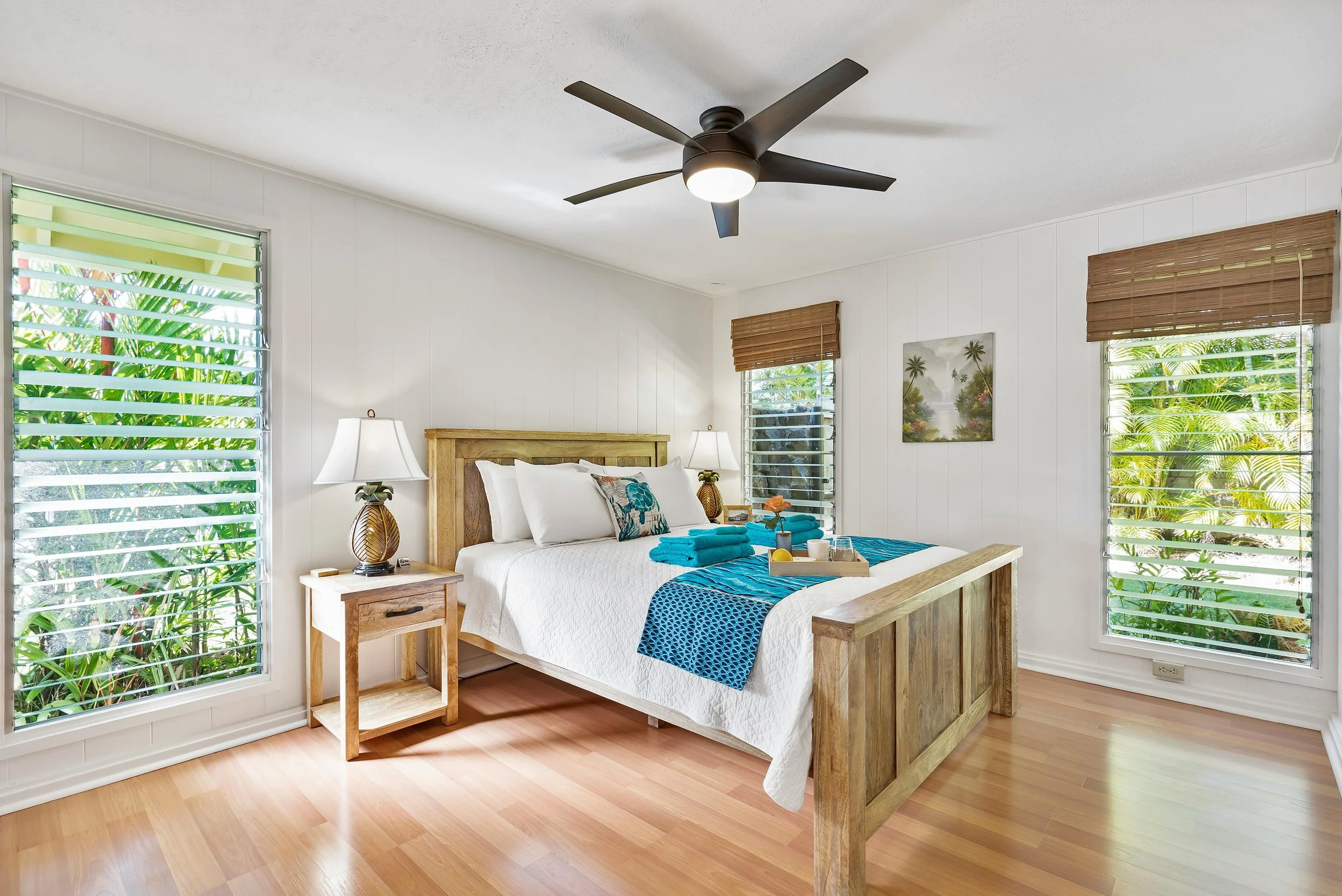 Bedroom with wooden bed frame, white bedding, and blue accents, two lamps, wooden nightstands, windows with blinds, tropical outdoor view, ceiling fan, light-colored wood flooring.