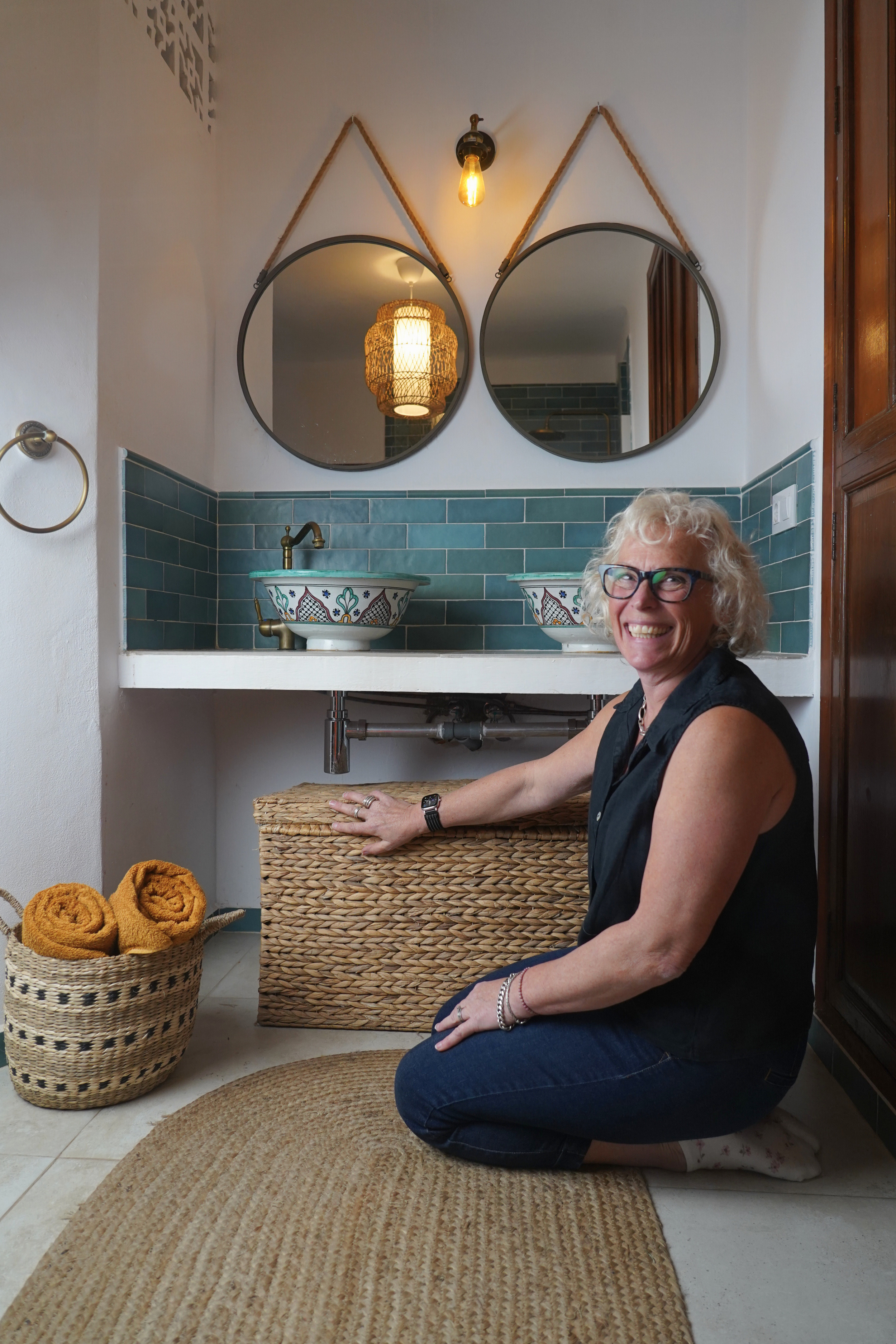Smiling woman with glasses kneeling on a bathroom floor, reaching towards a woven wicker basket, with two decorated vessel sinks and round mirrors behind her.