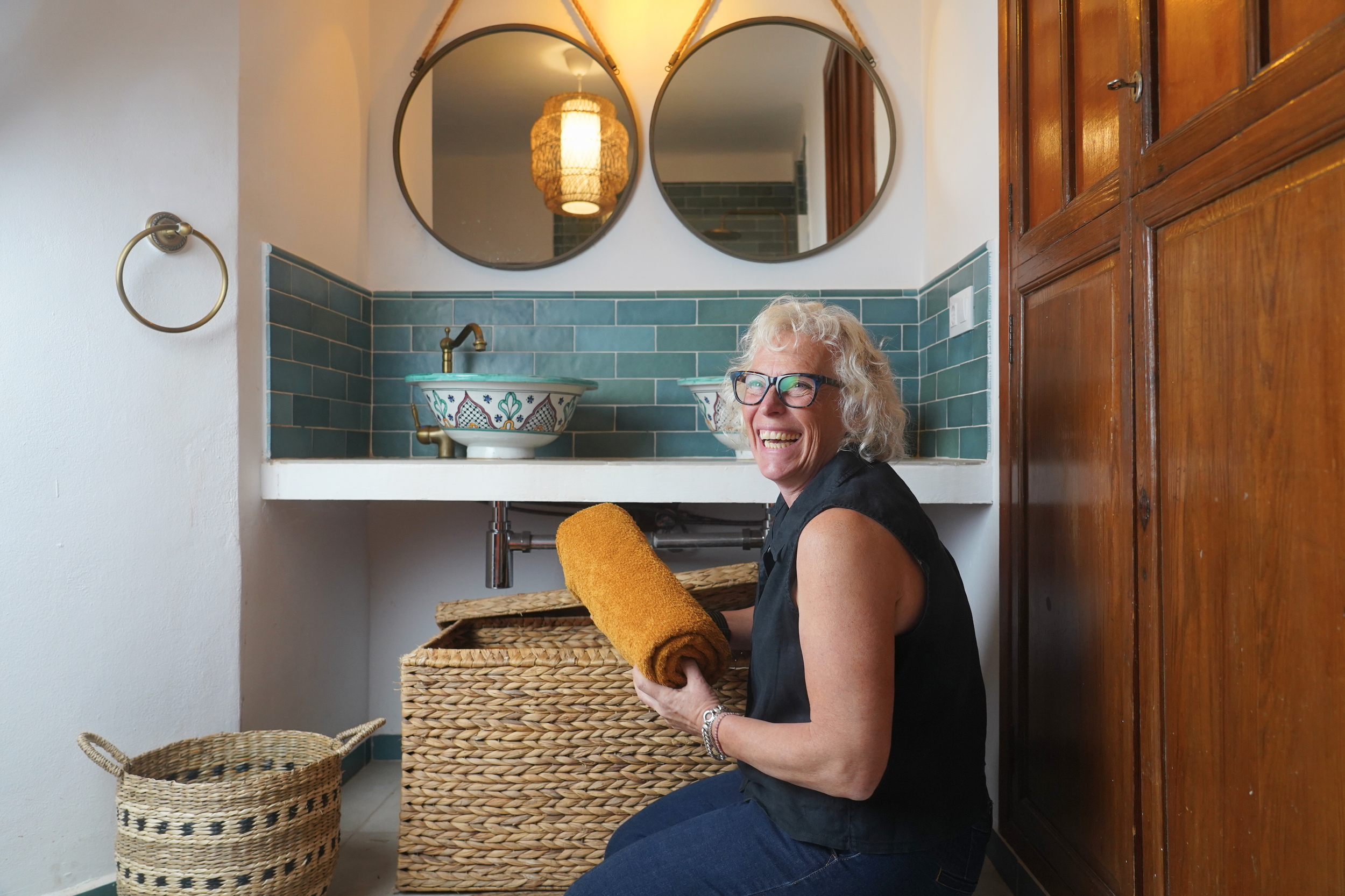 Woman smiling in a bathroom, holding a rolled yellow towel, kneeling beside a wicker laundry basket, with a bathroom sink with decorative bowls, mirrors, and wooden cabinet in the background.
