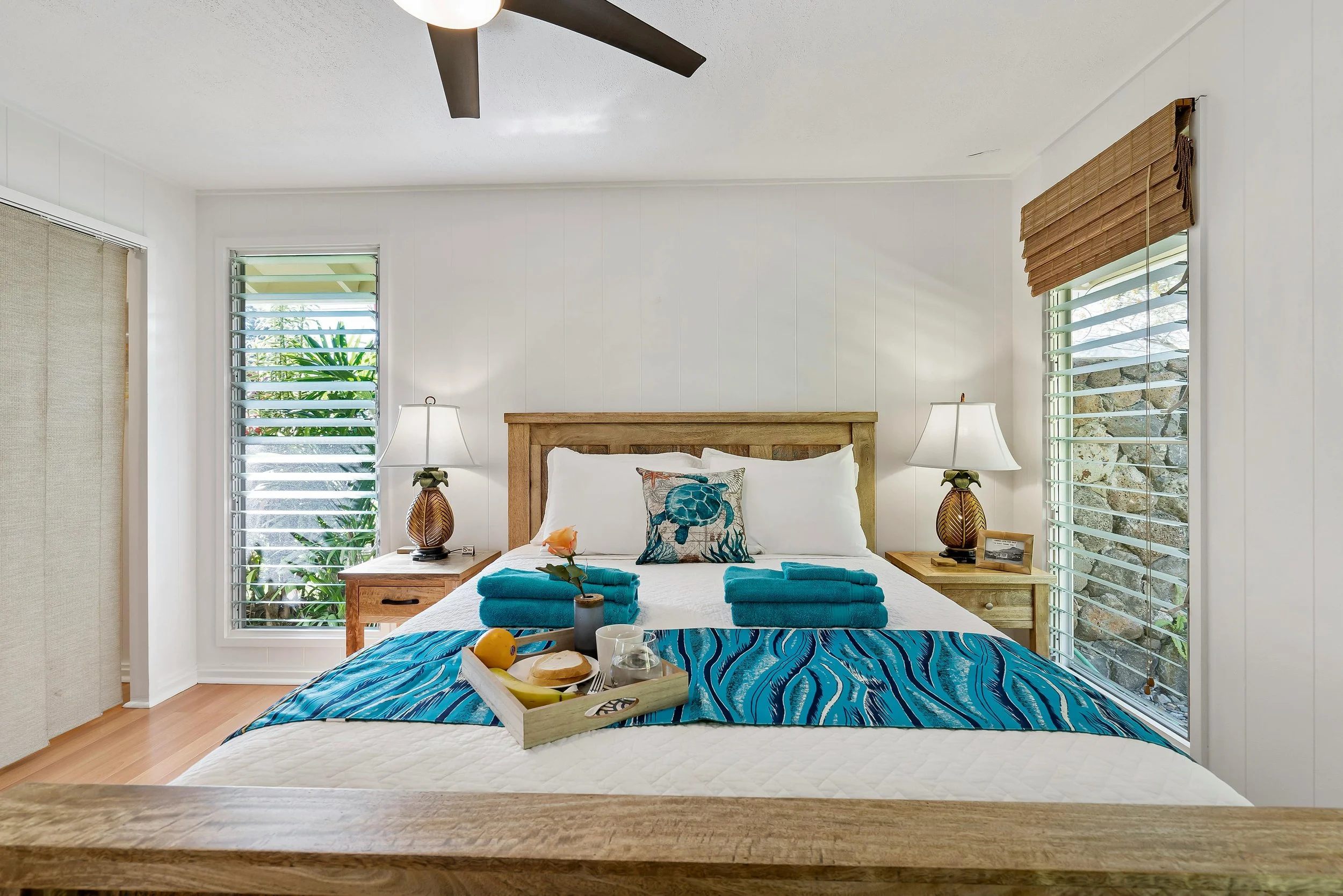 Bright bedroom with white walls, wooden bed frame, and two nightstands with lamps. The bed has white bedding, a decorative pillow with a turtle, and a blue patterned blanket. There are two teal towels and a tray with breakfast items on the bed. Large windows with shutters are on each side of the bed, letting in natural light.