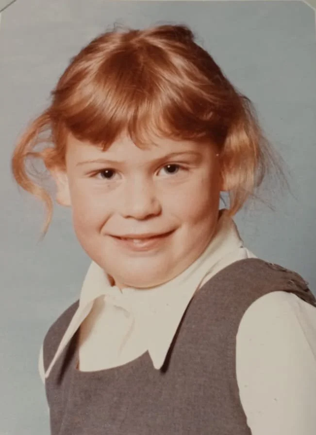 A young girl with red hair smiling and wearing a school uniform, including a white shirt and gray vest, against a plain background.