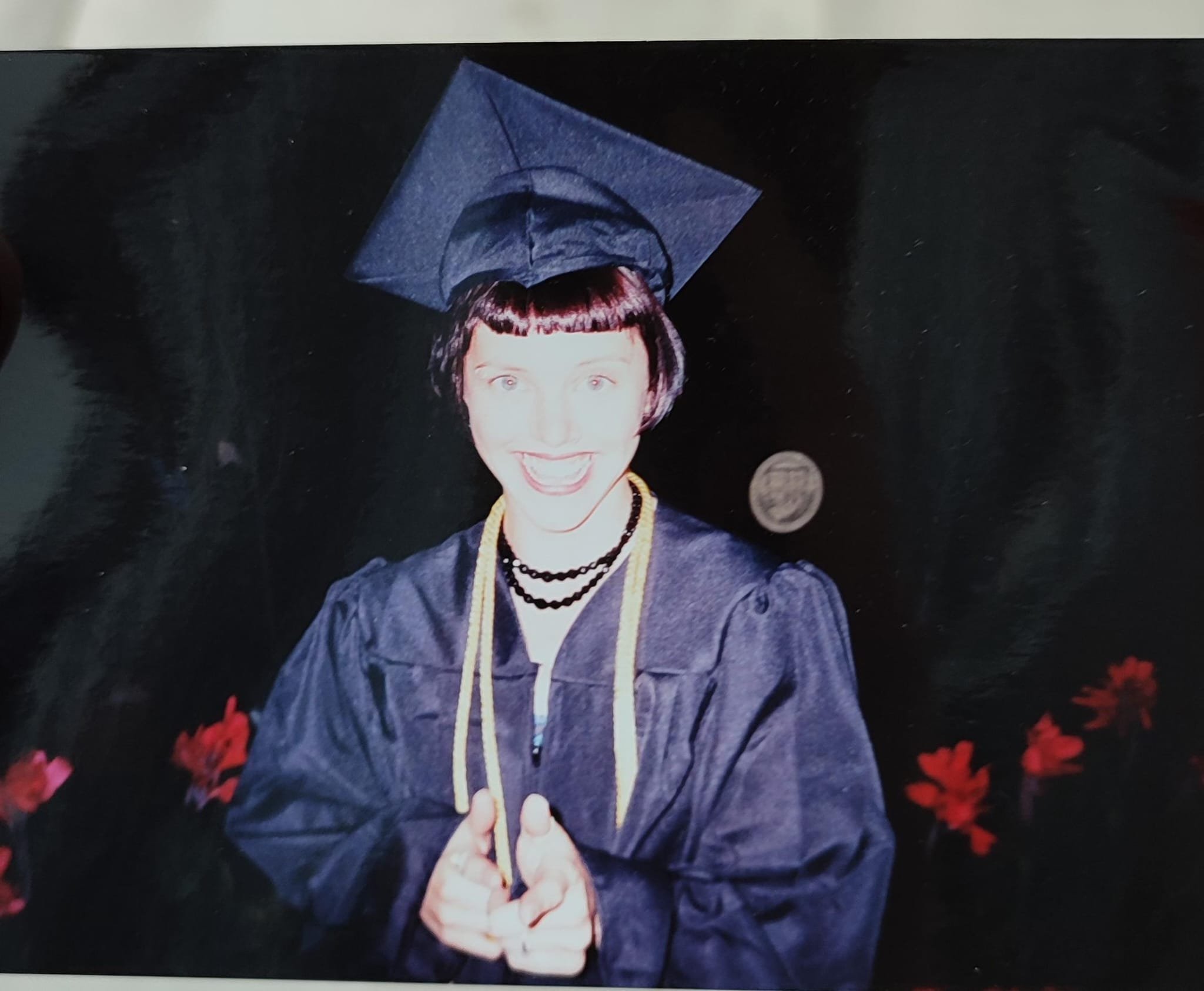 A woman in a graduation cap and gown smiling and pointing at the camera, with flowers in the background.