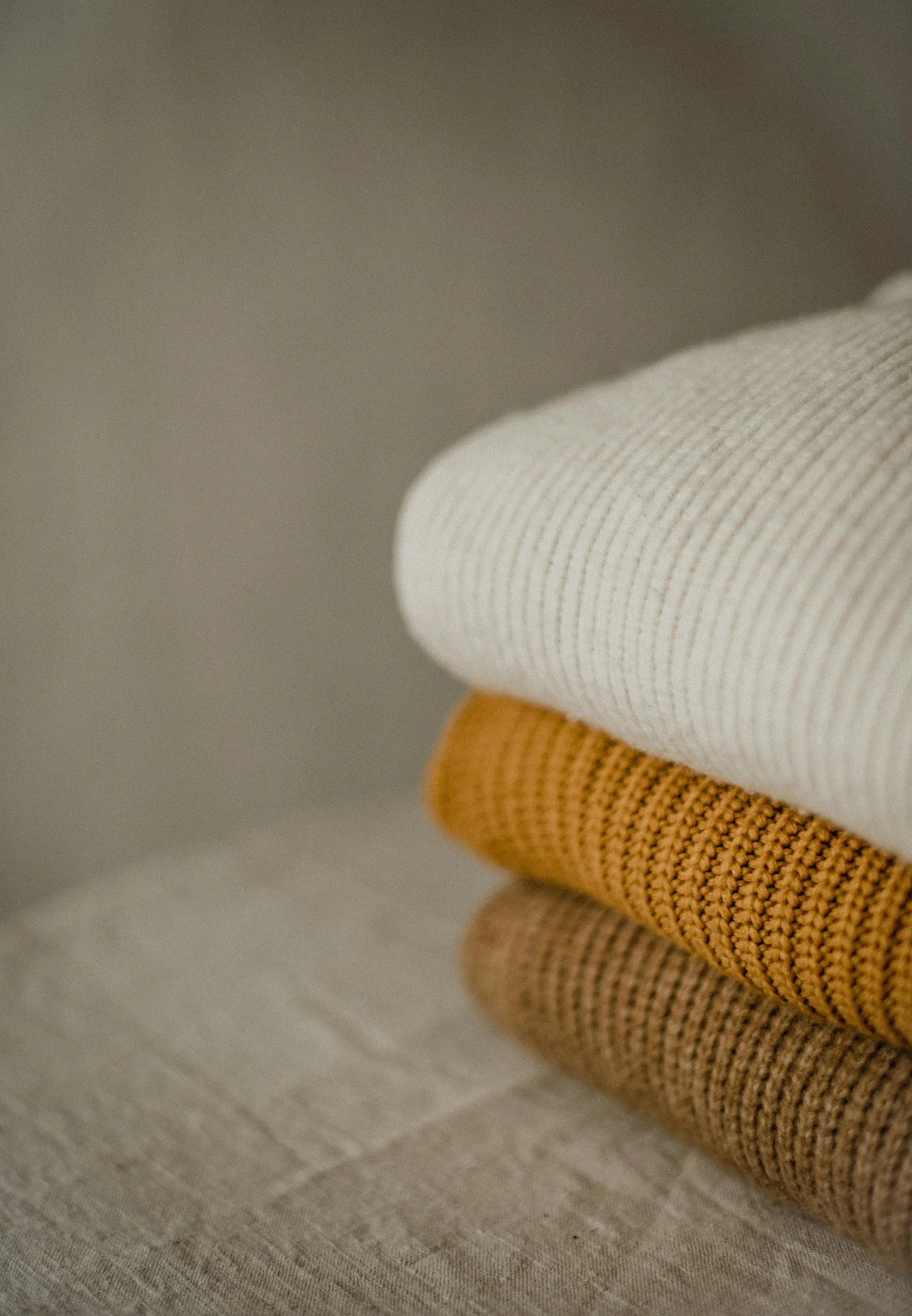 Stacked folded fabric or clothing in neutral colors, including white, mustard, beige, and light brown.