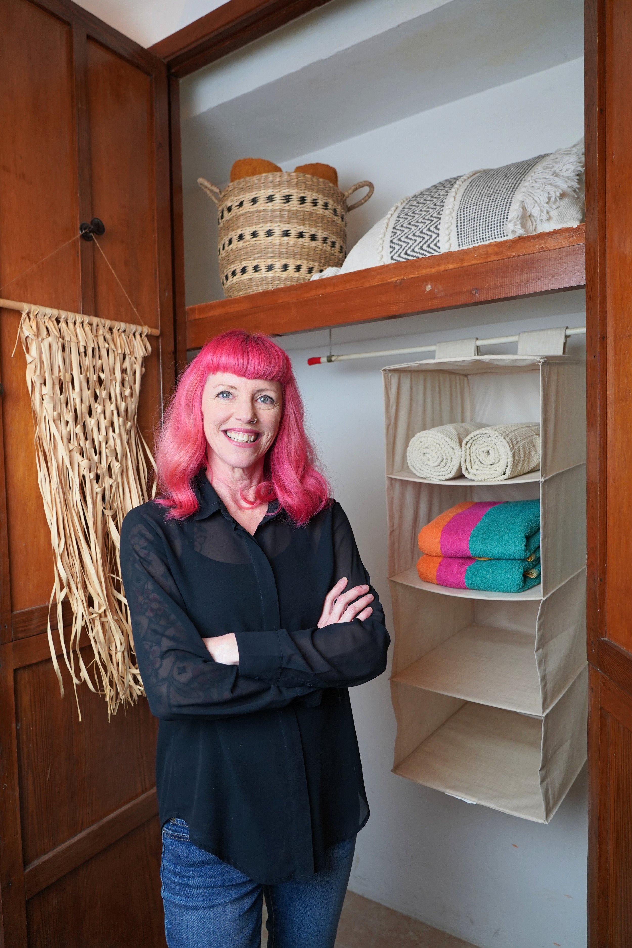 A woman with pink hair and a black blouse standing with arms crossed in a linen closet filled with towels and storage baskets.