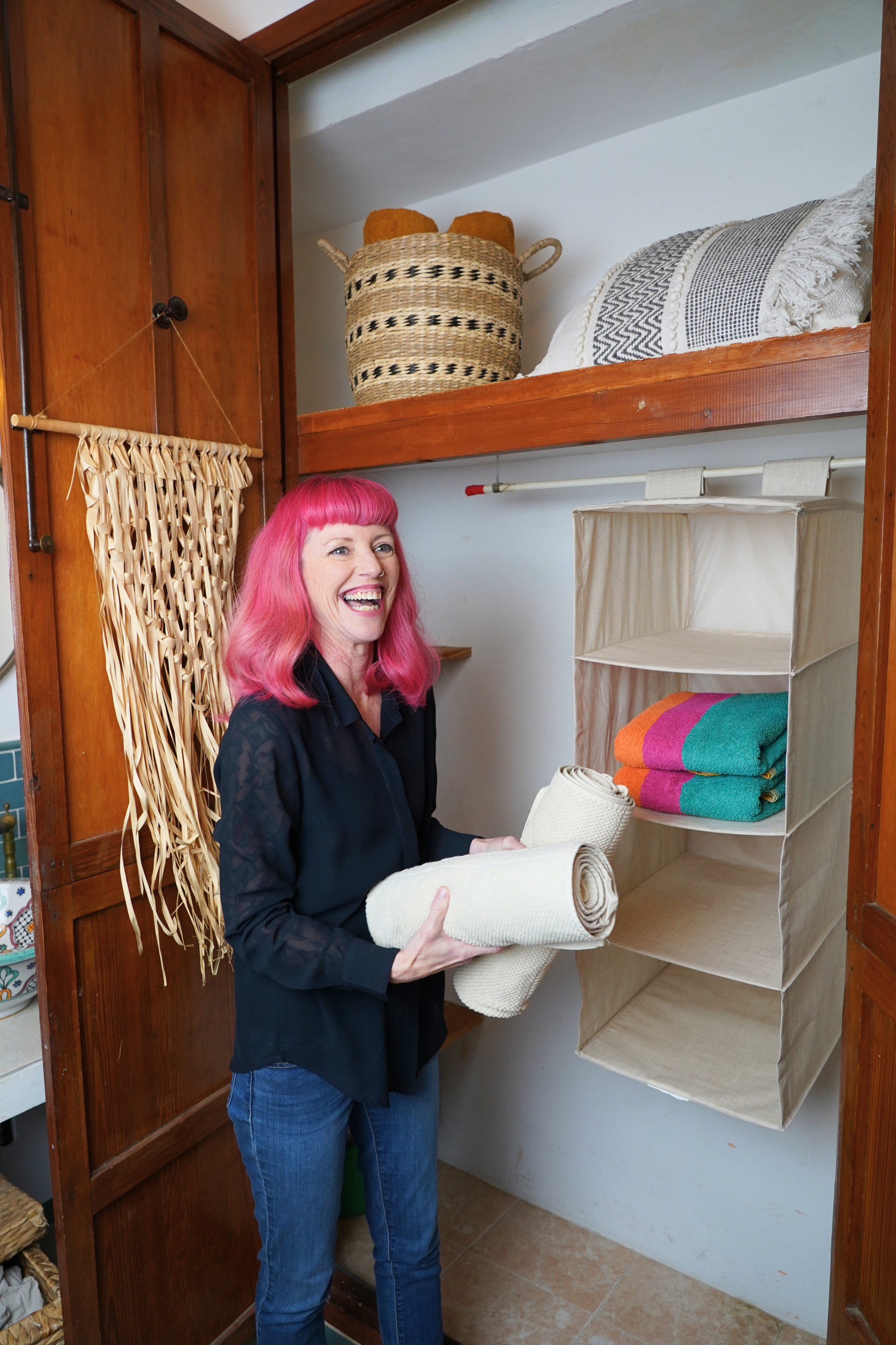 Woman with pink hair smiling while holding rolled towels in a closet with shelves containing colorful towels and baskets.