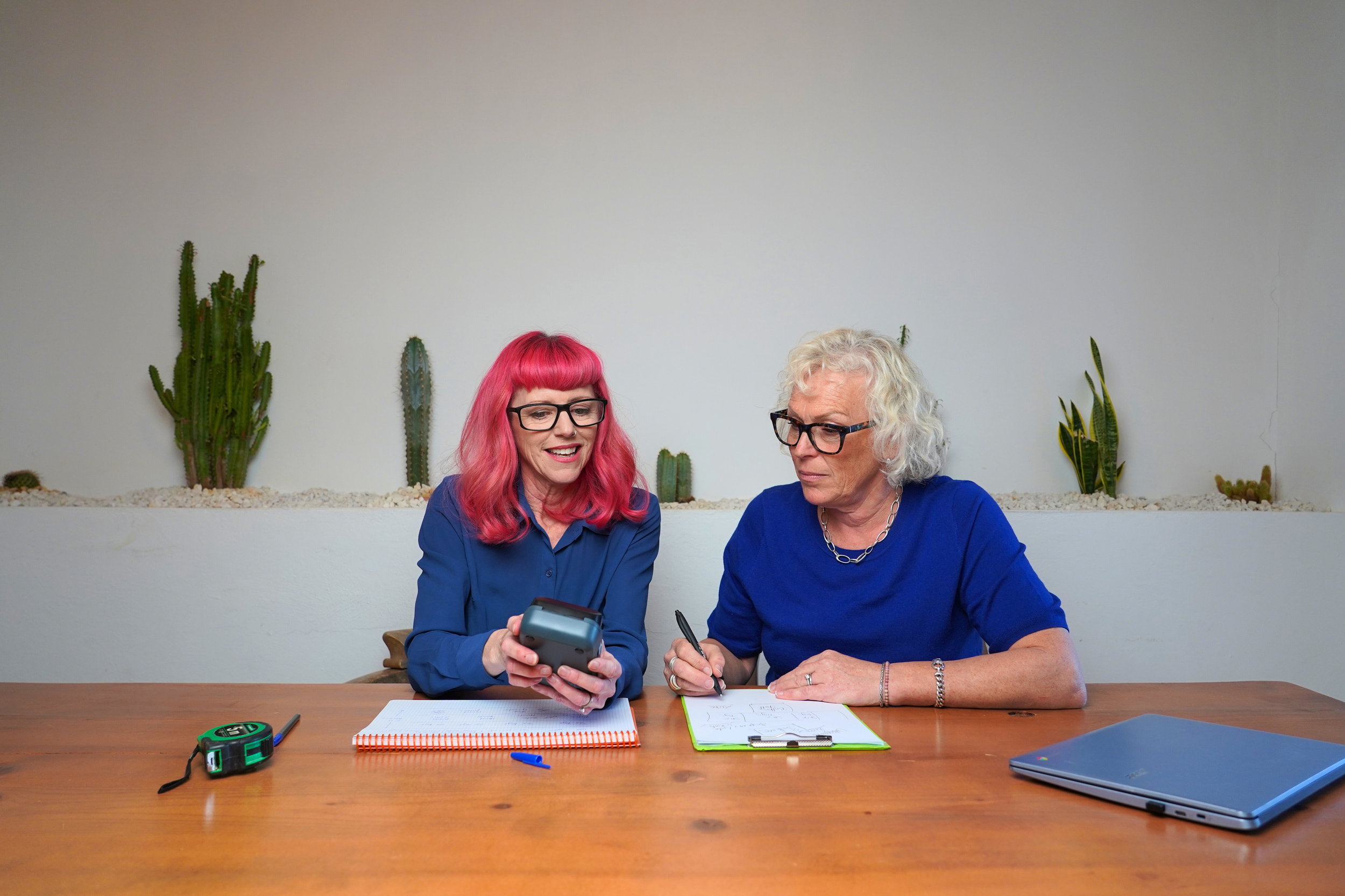 Two women sitting at a wooden table with notebooks, a laptop, and a tape measure, looking at a smartphone together, with cacti and succulents in the background.