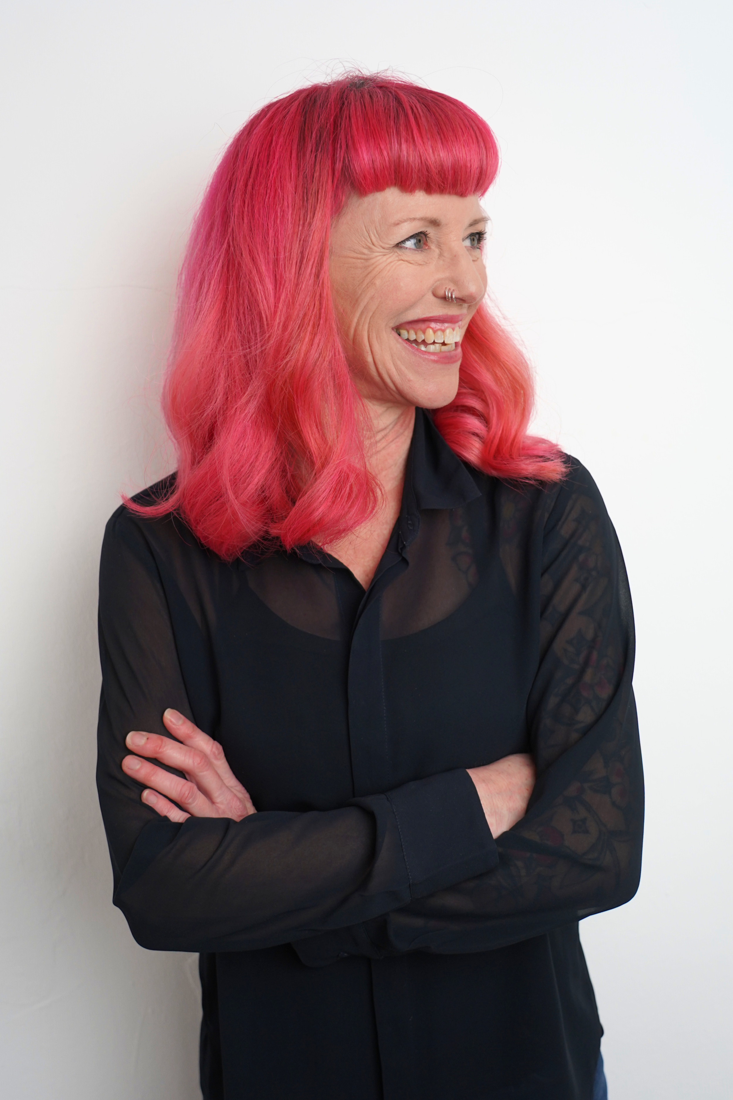 A woman with bright pink hair and a nose ring, wearing a black, sheer long-sleeved shirt, smiling and crossing her arms against a white background.