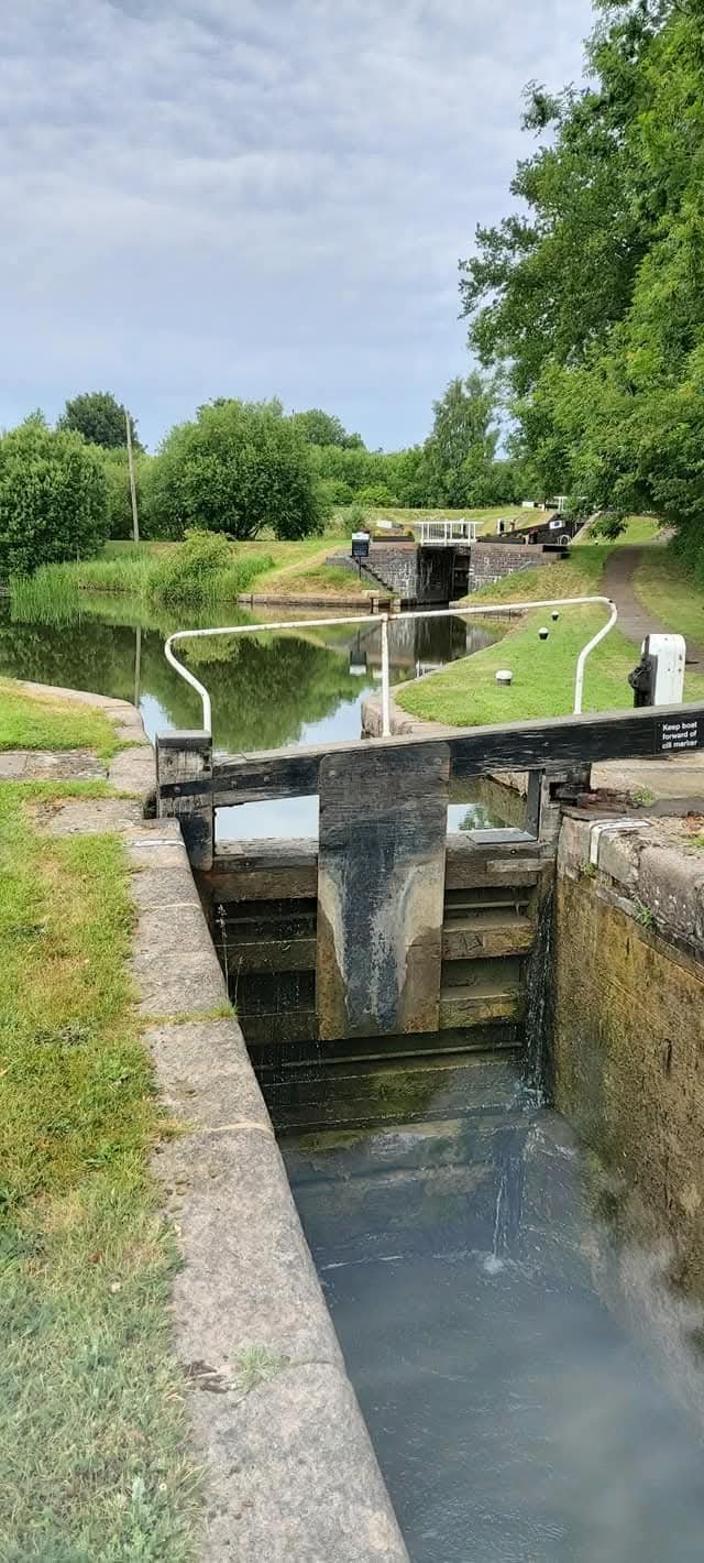 A canal lock system with a water lock gate, grassy pathway, and surrounding lush trees, under a partly cloudy sky.
