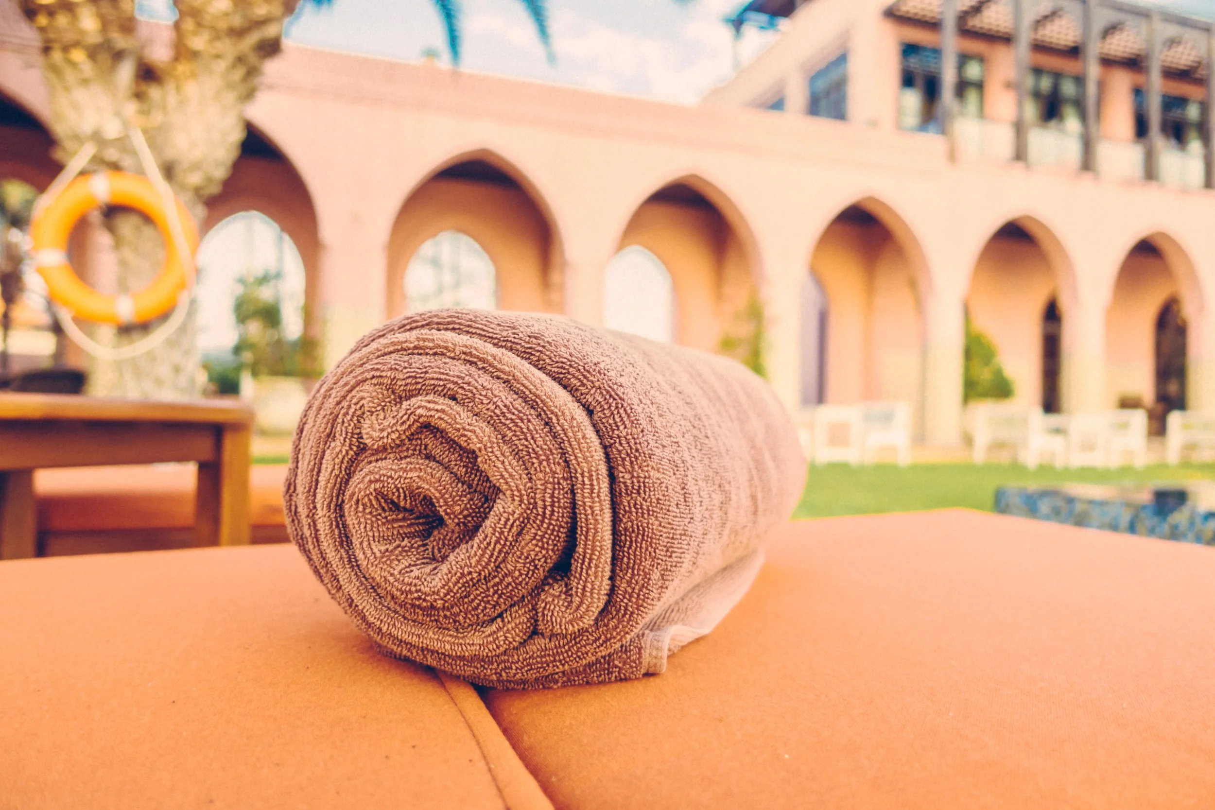 Rolled brown towel placed on an orange outdoor lounge chair with a courtyard, grass, white chairs, and an arched building in the background.