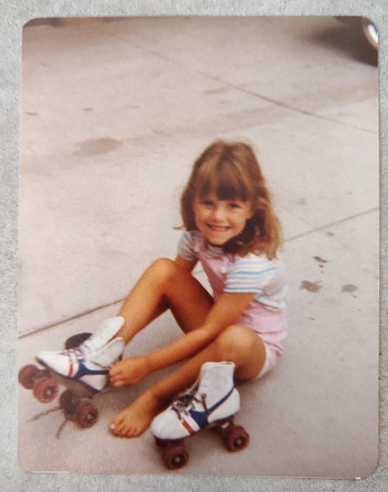 A young girl with brown hair is sitting on the pavement on a skateboard with her legs crossed, smiling at the camera. She is wearing a striped shirt, pink shorts, and white sneakers.