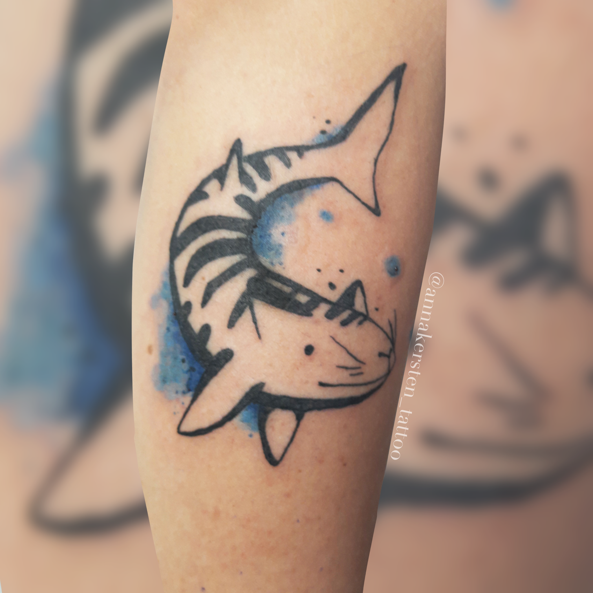 Tätowierung eines Haifisches in einem schwarz-blauen Design auf der Haut, mit bläulichen Aquarell-Flecken im Hintergrund.