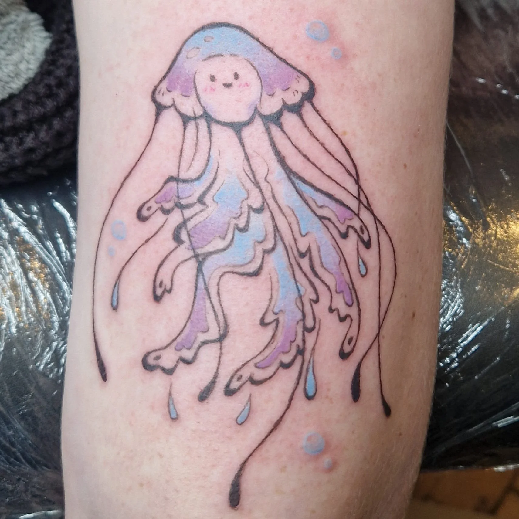 Tätowierung eines niedlichen, cartoonartigen Quallenmotivs auf der Haut, mit blauen und lilafarbenen Akzenten.