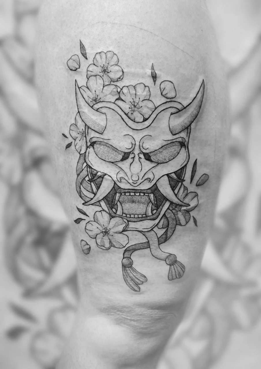 Schwarze Tätowierung eines dämonischen Gesichts mit Hörnern, Blumen und Ranken auf der Haut.