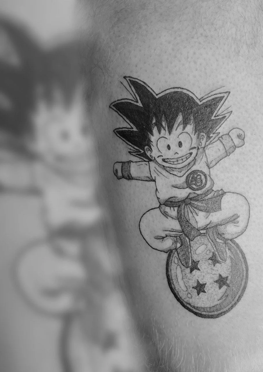 Schwarz-weiß-Tattoo eines Jungen mit Goku-Haare auf einem Ball. Der Junge lächelt, trägt ein T-Shirt mit einem Symbol und einen Gürtel, und sitzt auf einem Ball mit Sternen.