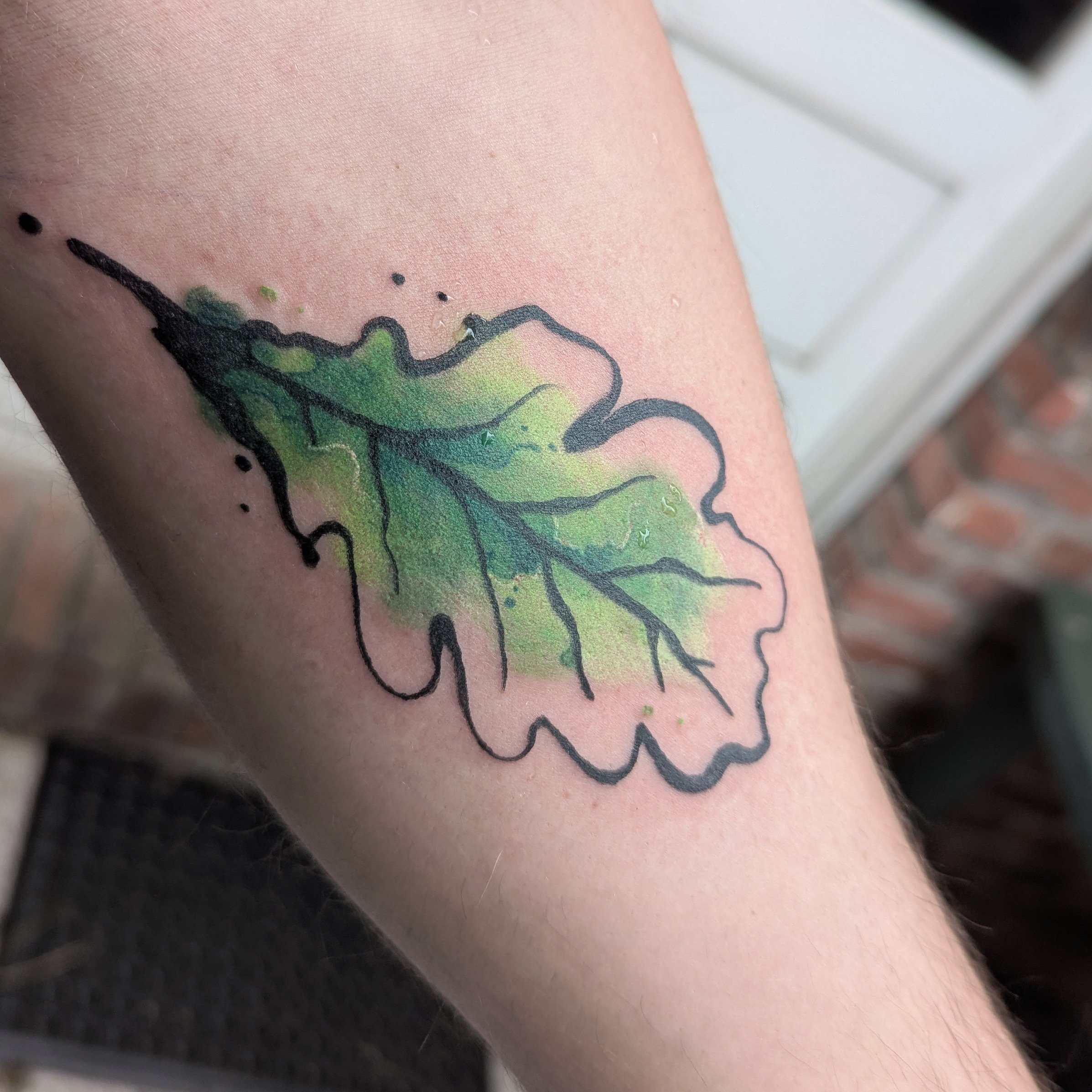 Tattoo von einem grünen Eichenblatt mit schwarzem Umriss auf dem Arm.