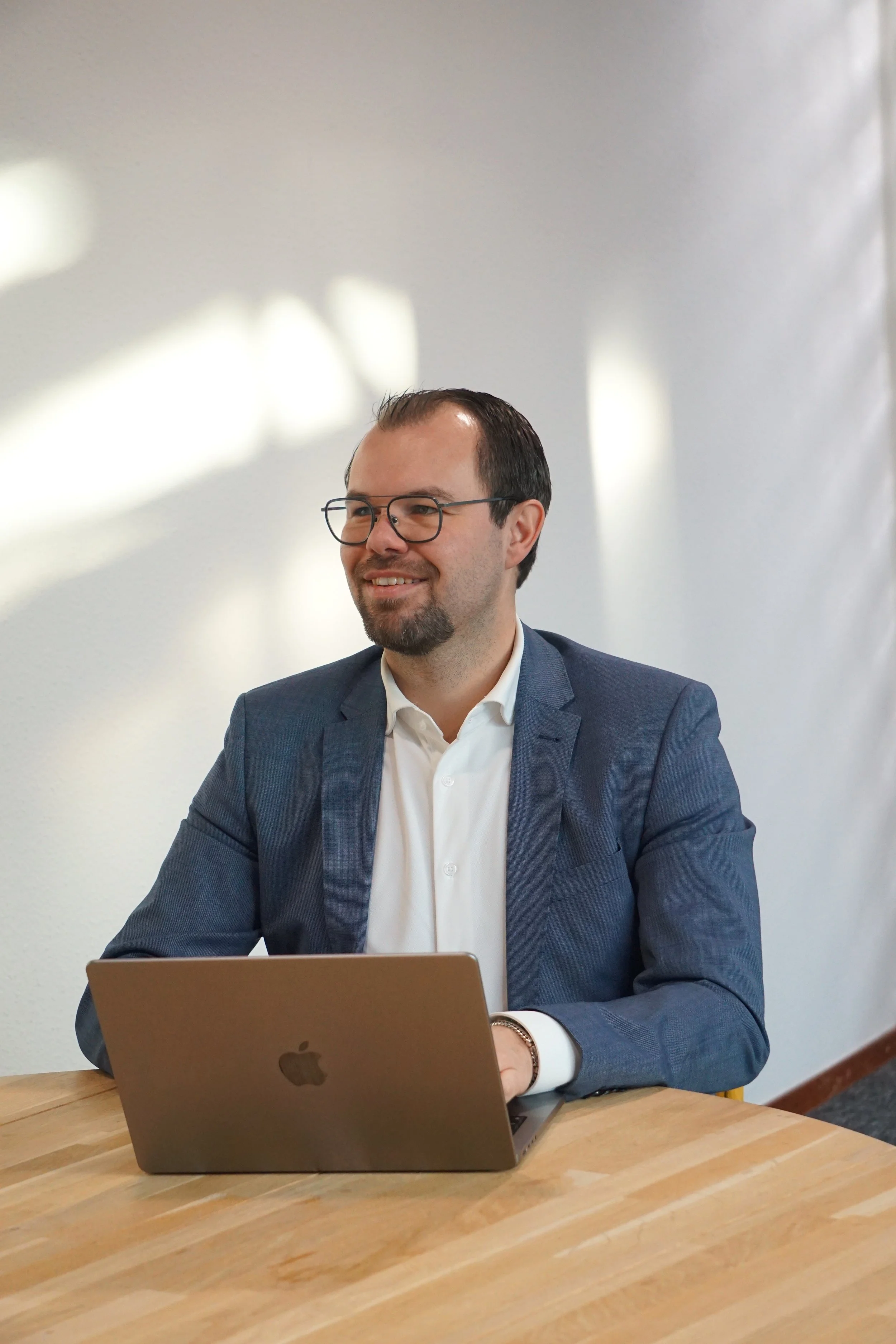 Man in een blauw pak met bril zit aan een houten tafel, werkt op een zilveren MacBook, op de achtergrond een witte muur met lichte schaduwen.