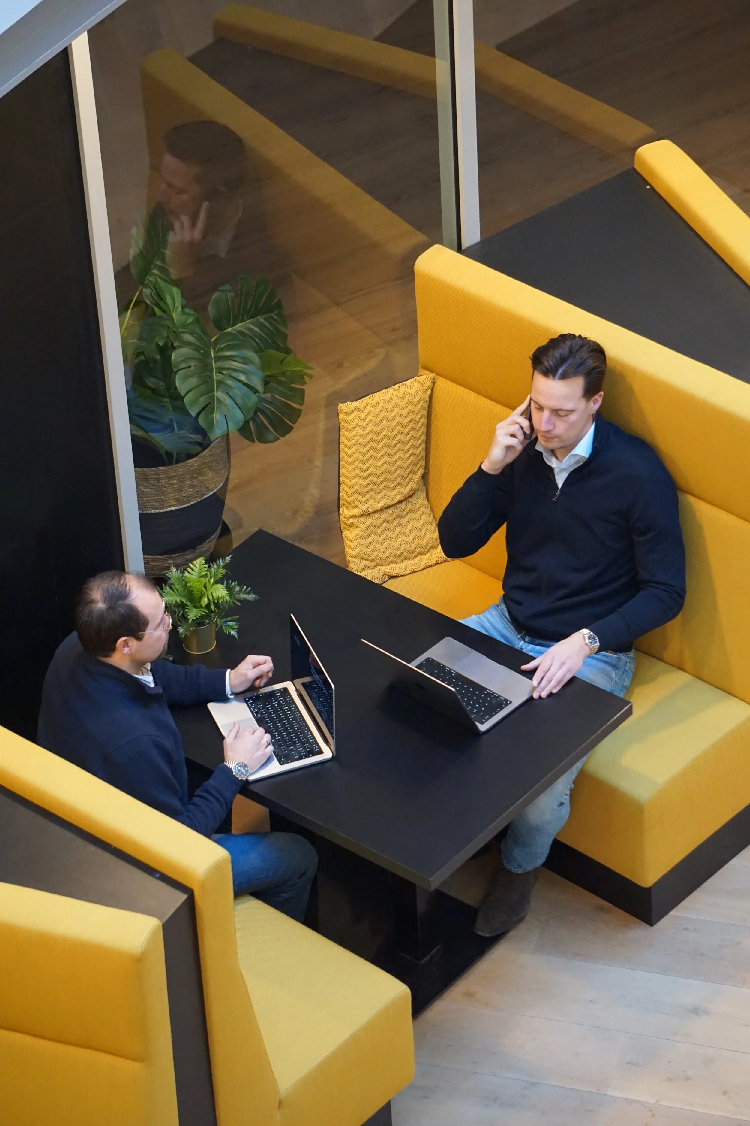 Twee mannen werken met laptops in een booth met gele bekleding en grote planten.