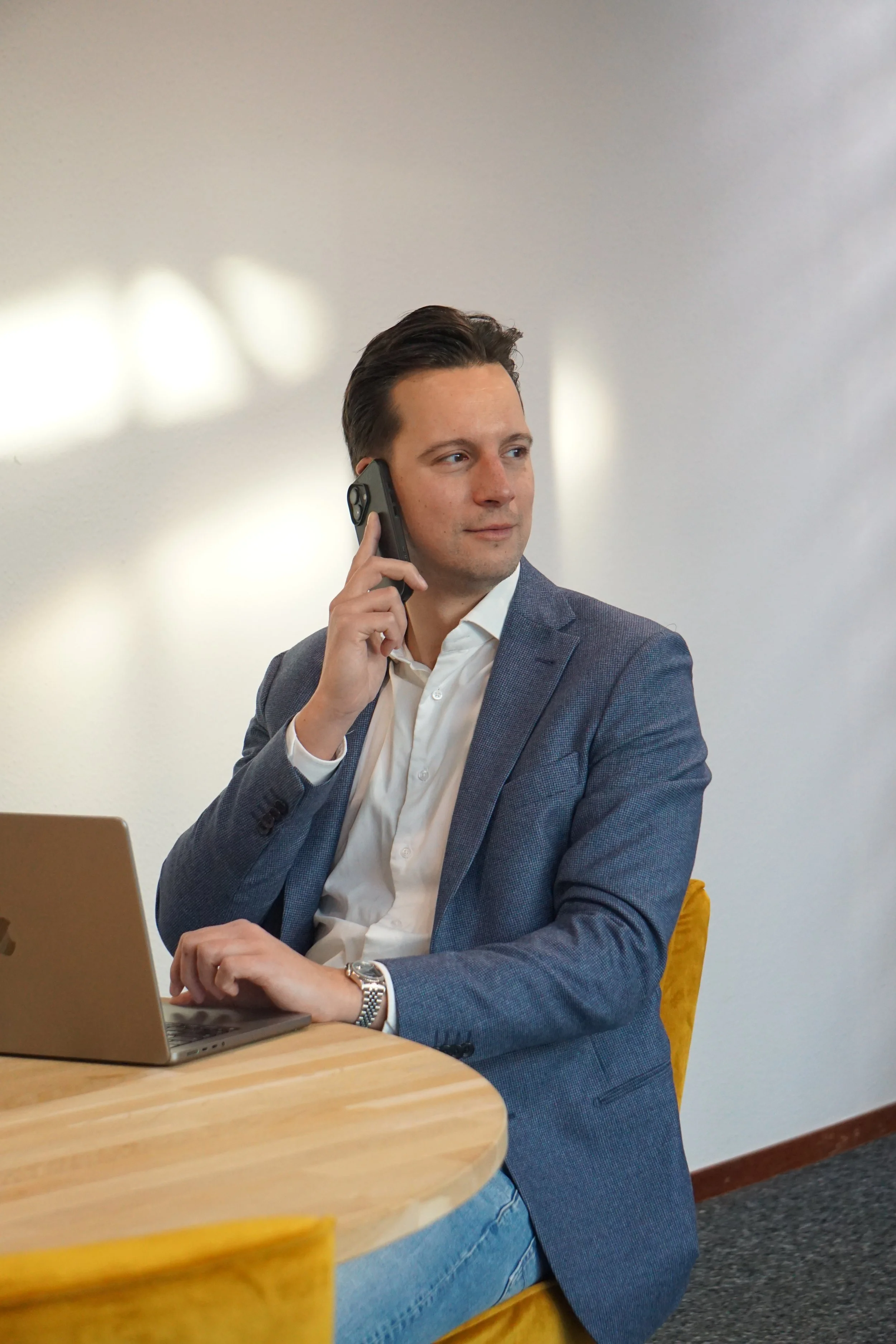 Man in een grijze jas en witte blouse zit aan een houten tafel, belt met zijn mobiele telefoon en heeft een laptop voor zich op de tafel.