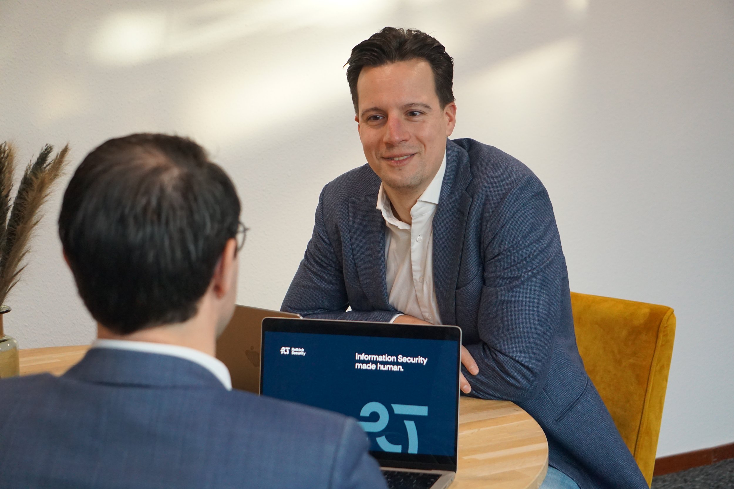 Twee mannen hebben een gesprek op een kantoor. Ze zitten aan een tafel met laptops voor zich. Een van hen gebruikt een laptop met een scherm dat een zakelijke presentatie toont.