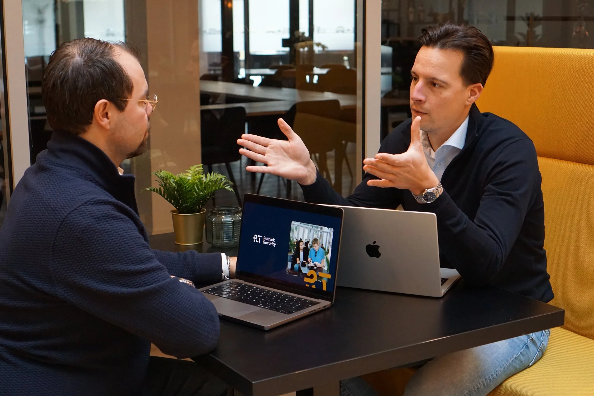 Twee mannen in gesprek in een kantoorruimte, één toont met handgebaar terwijl de ander luistert, op een tafel met laptops en een plant.