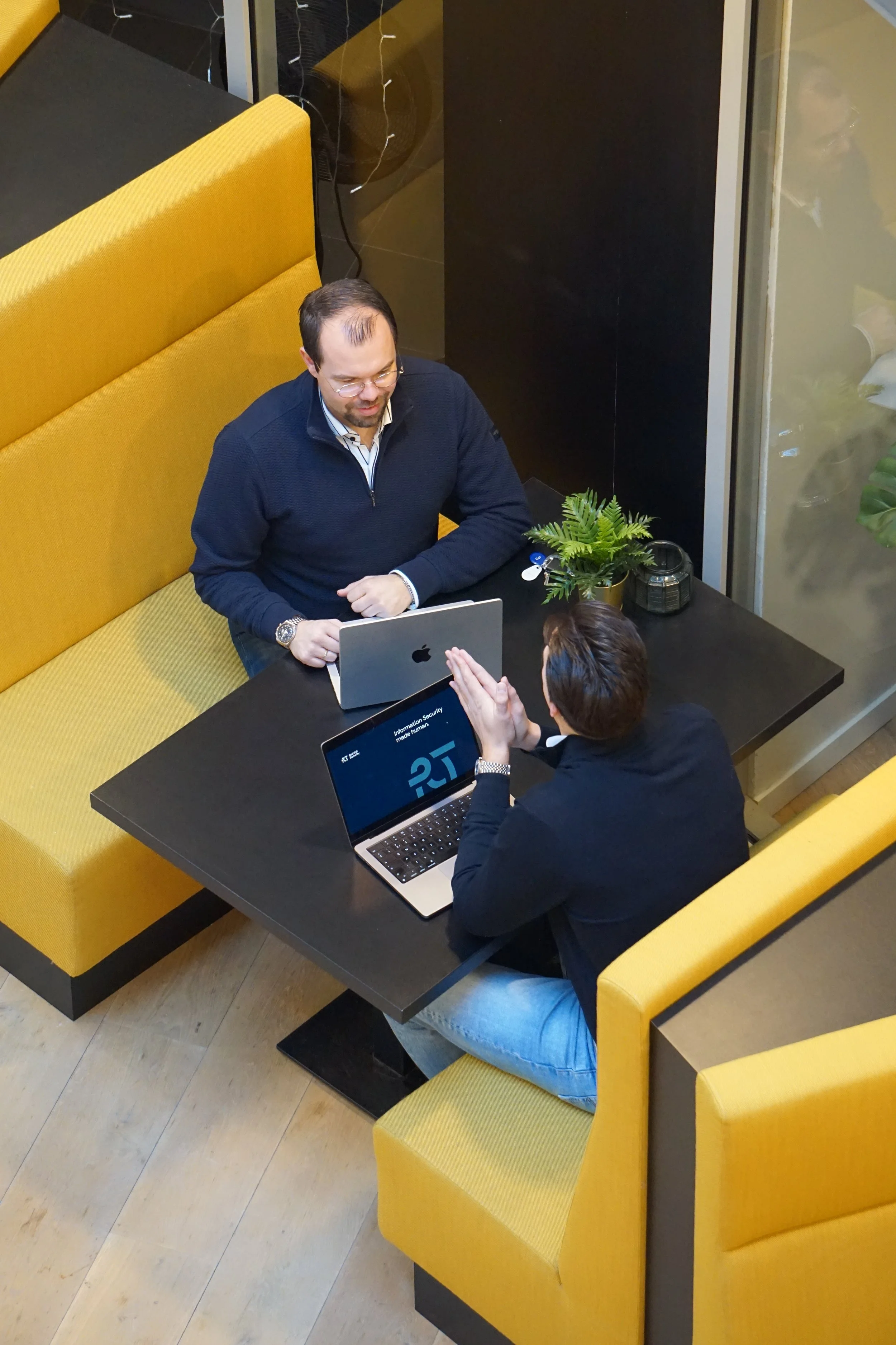 Twee mensen zitten aan een zwarte tafel in een restaurant, met gele banken op de achtergrond. Ze praten terwijl ze met elkaar en laptops werken, en er staat een plant op de tafel.