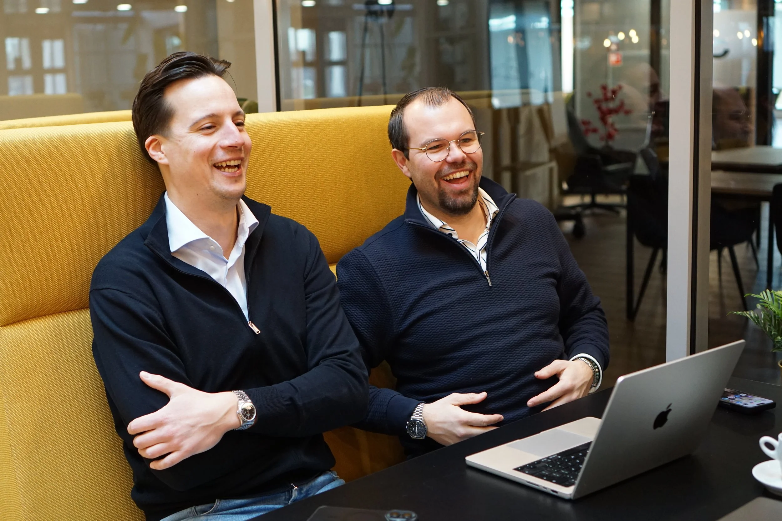 Twee mannen lachen terwijl ze achter een laptop zitten in een moderne kantoorruimte.