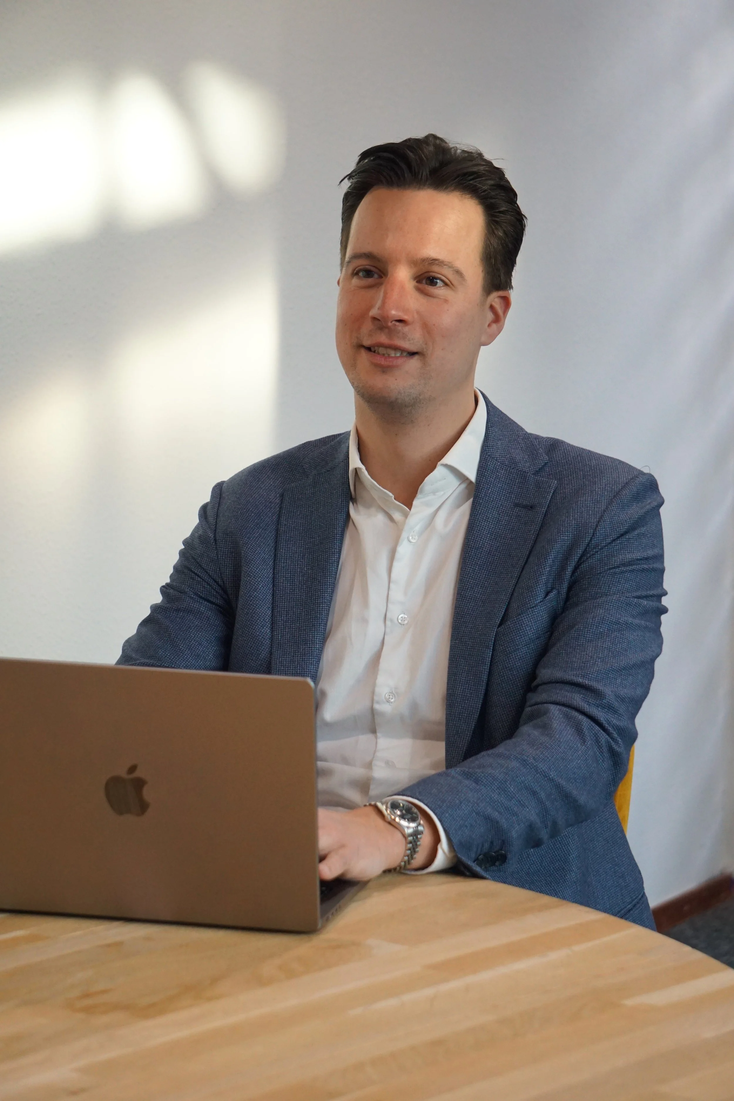 Man in een grijs pak en witte overhemd zit aan een houten tafel met een open laptop voor hem, achtergrond is effen lichtgrijs.