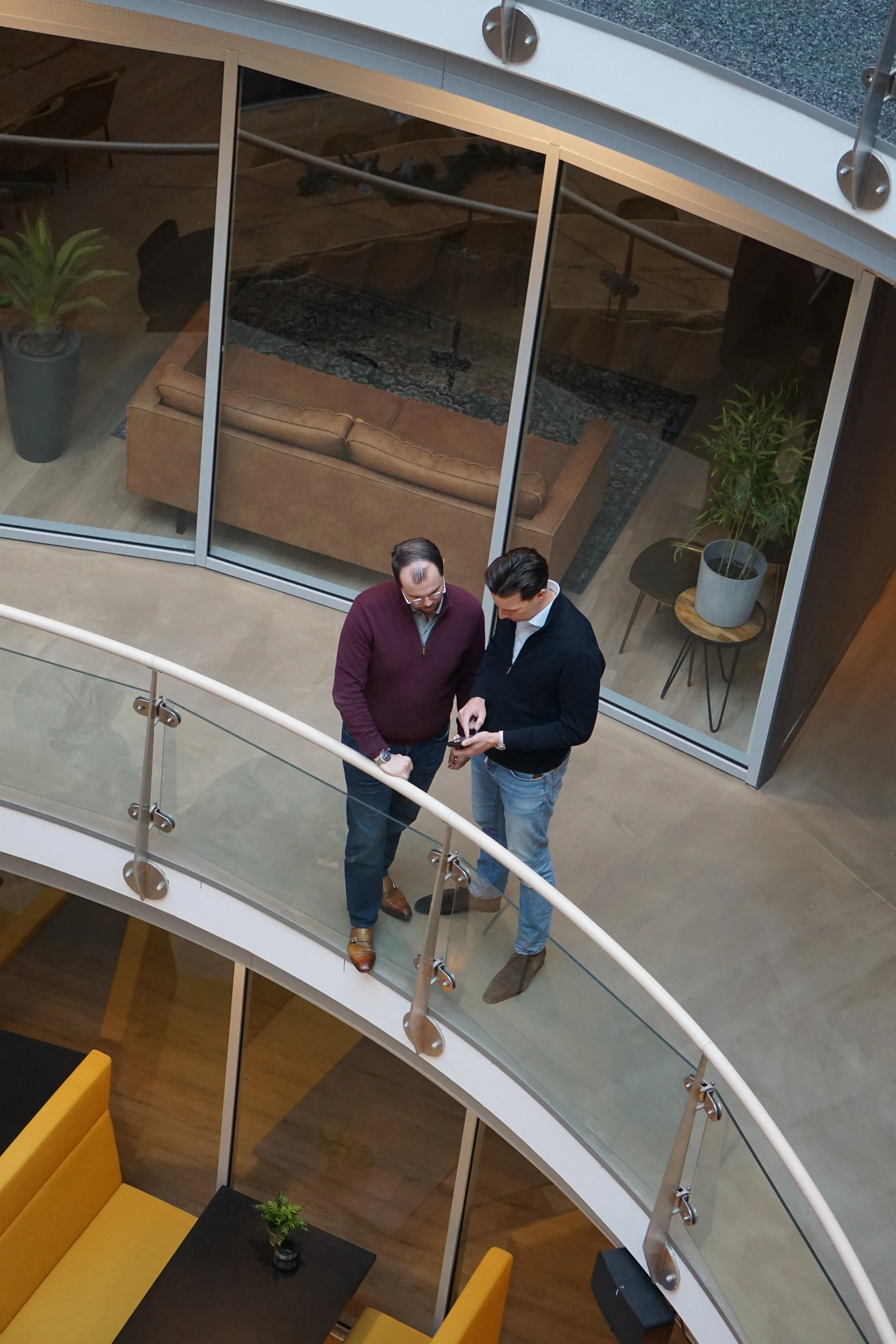 Twee mannen die samen een smartphone bekijken op een balkon van een kantoor of hotel, met een glazen muur en binnenruimte met een bank en planten erachter.