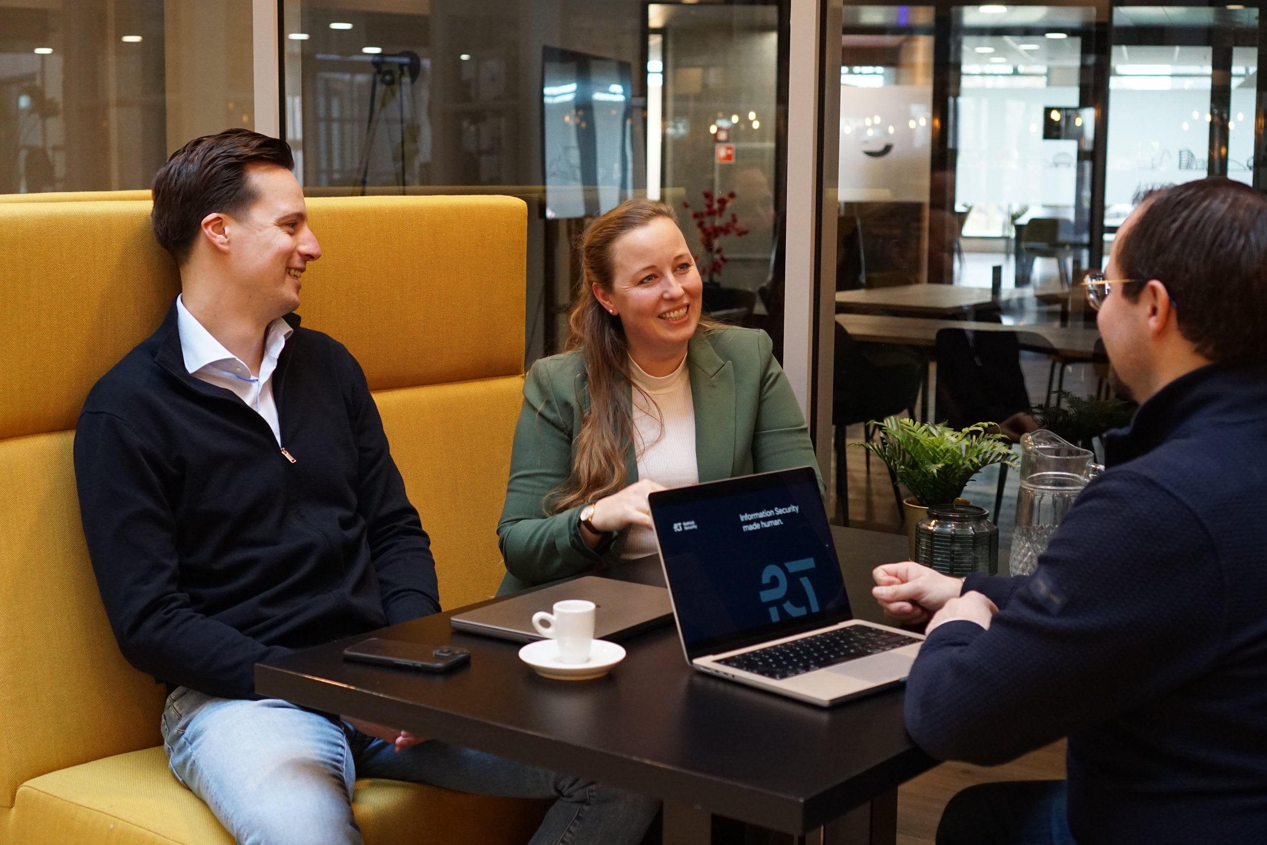Drie mensen die vrolijk met elkaar praten. Ze zitten aan een tafel met een laptop, een kopje espresso en een mobiele telefoon. De sfeer lijkt informeel en vriendelijk.