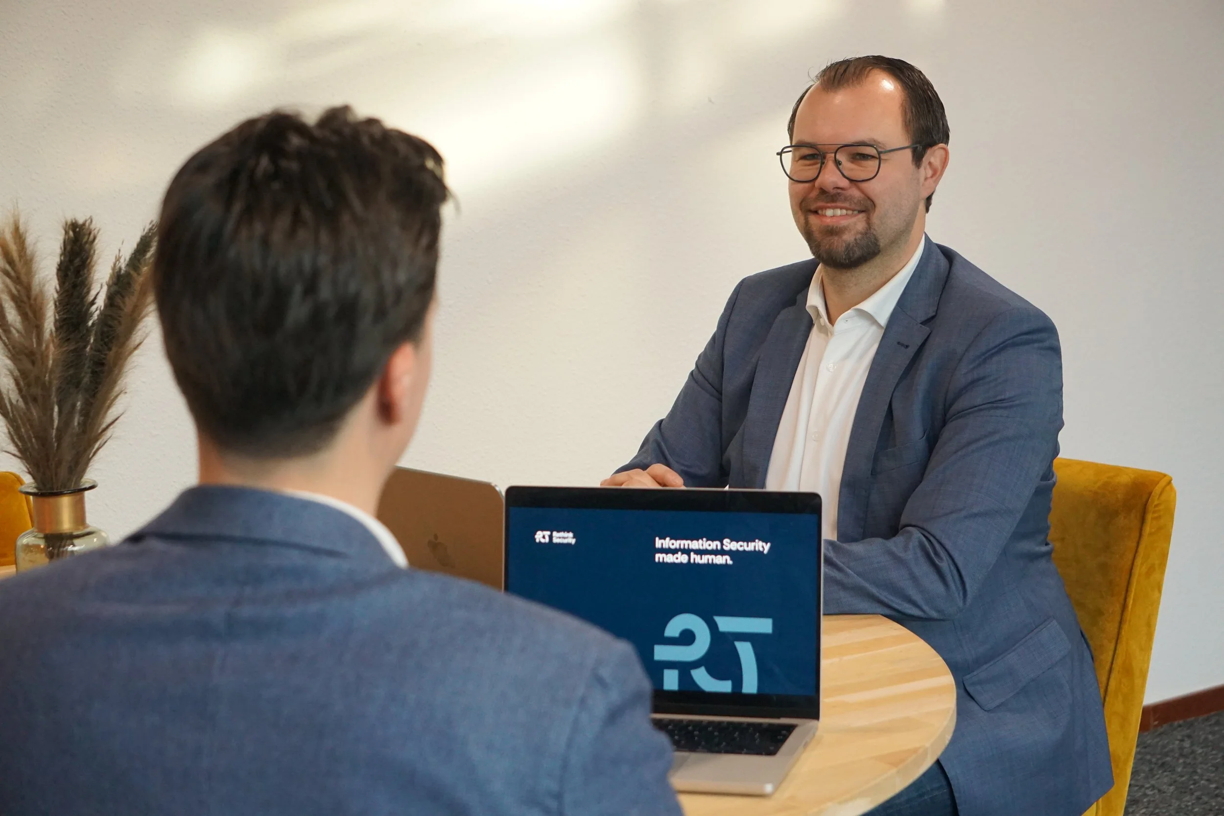 Twee mannen hebben een gesprek op een kantoor. Ze zitten aan een tafel met laptops voor zich. Een van hen gebruikt een laptop met een scherm dat een zakelijke presentatie toont.