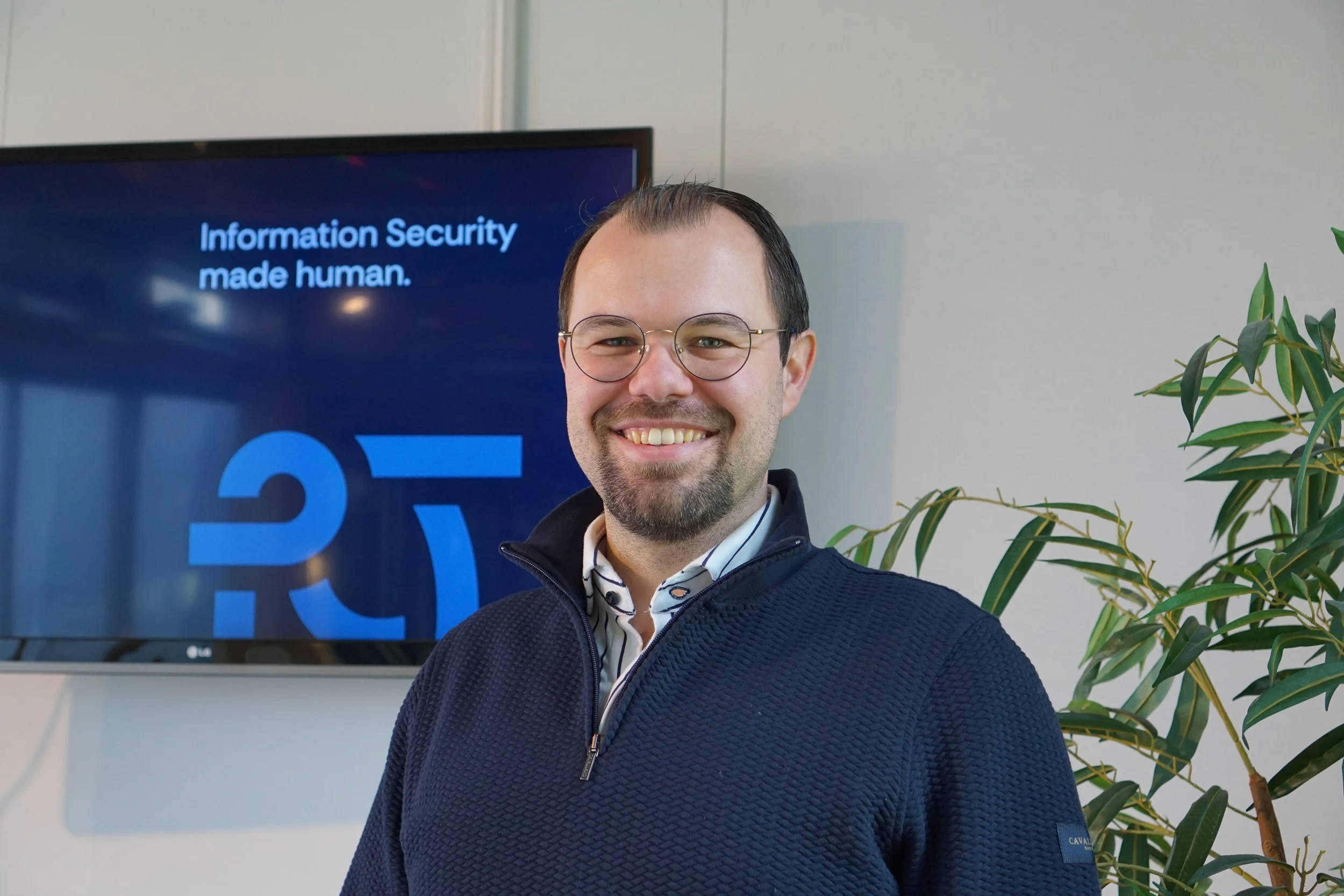 Man met bril, glimlach, gekleed in donkerblauw vest, kledingstijl met streepjes, staat voor een scherm met tekst 'Information Security made human' en een groene plant rechts.