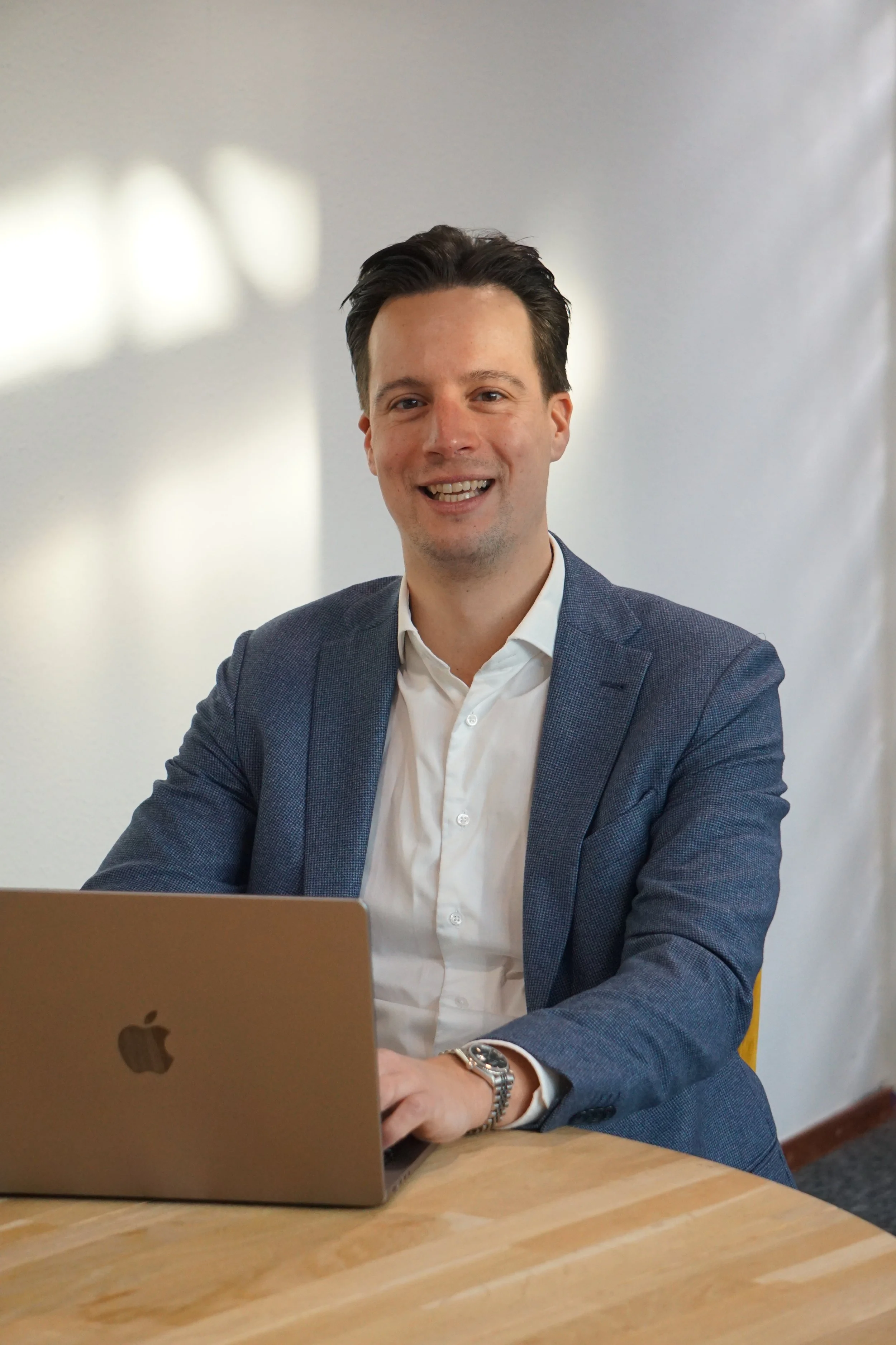 Man in een grijs pak werkt op een laptop, zittend aan een houten tafel.