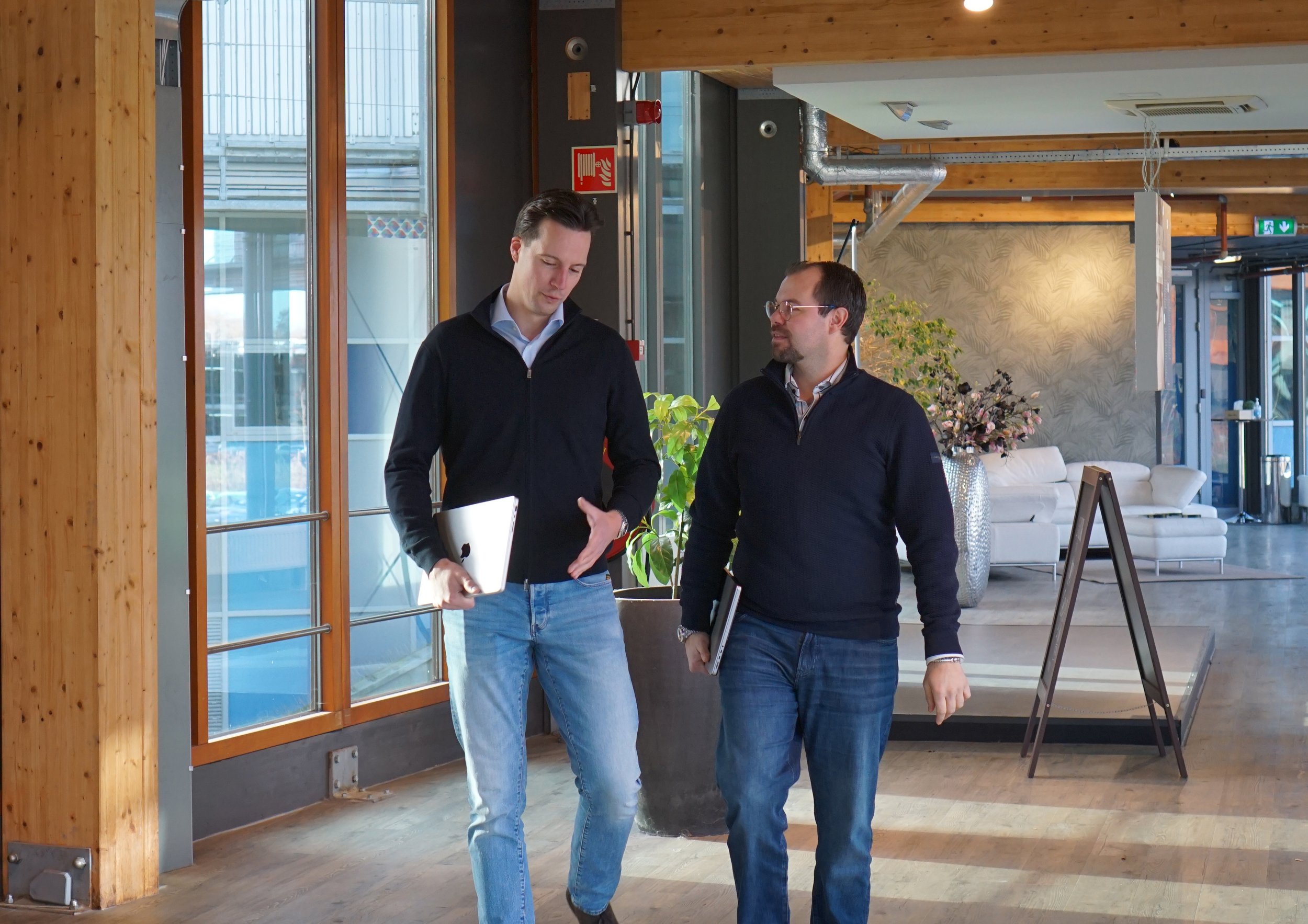 Twee mannen lopen door een moderne binnenruimte, één houdt een laptop onder zijn arm en ze voeren een gesprek.
