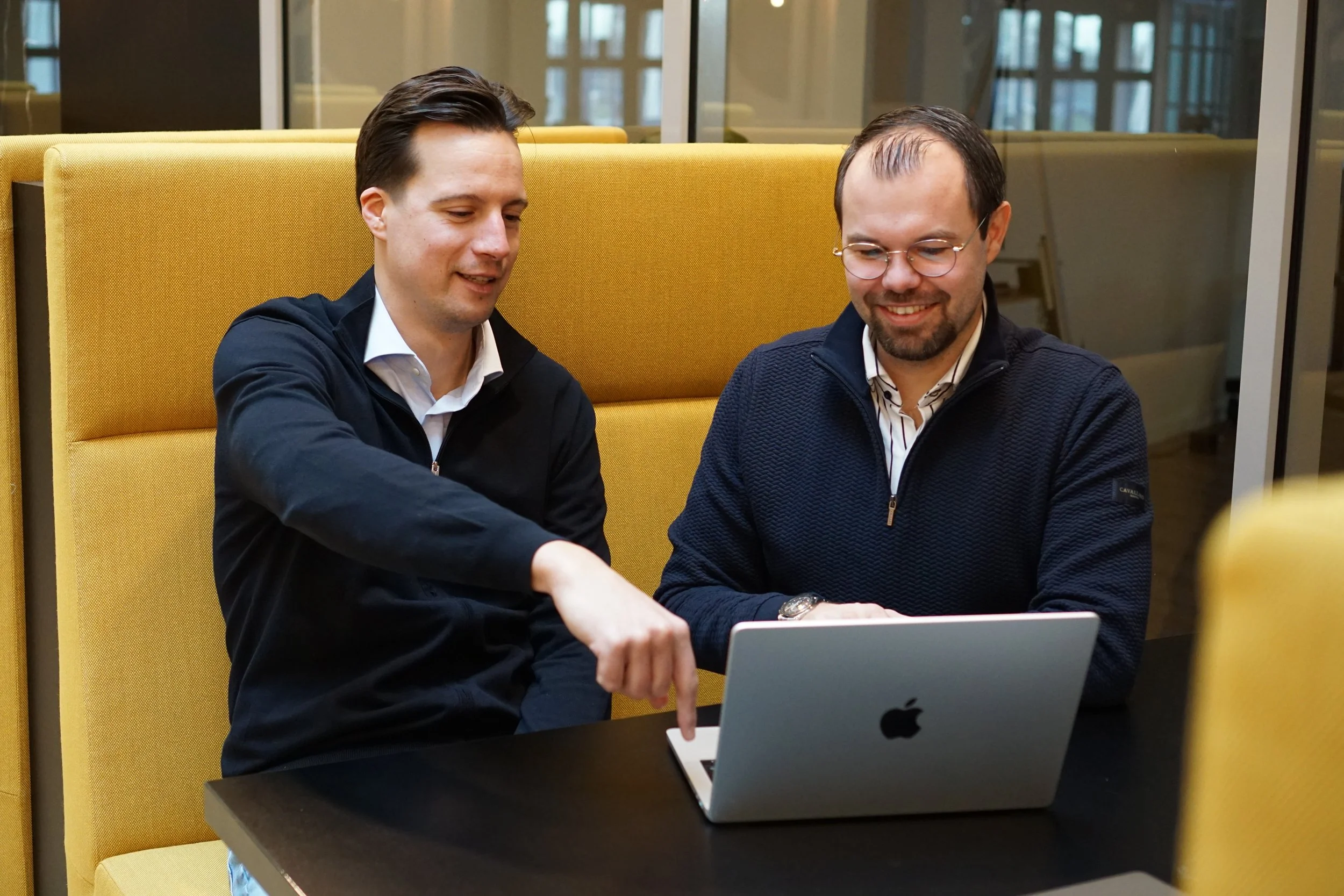 Twee mannen werken samen op een laptop in een kantooromgeving, ontspannen en glimlachend.