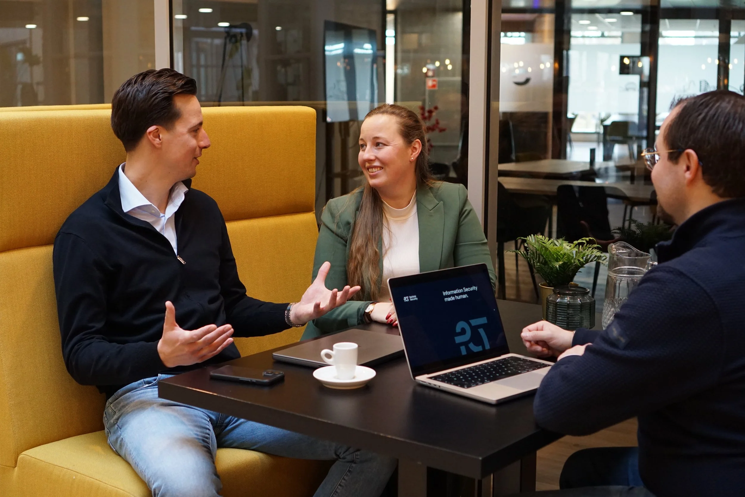 Drie mensen die in een vergaderzaal zitten, betrokken in een gesprek. Een man links praat, terwijl een vrouw en een man rechts luisteren. Op de tafel staan een laptop, een kopje koffie en een plant.