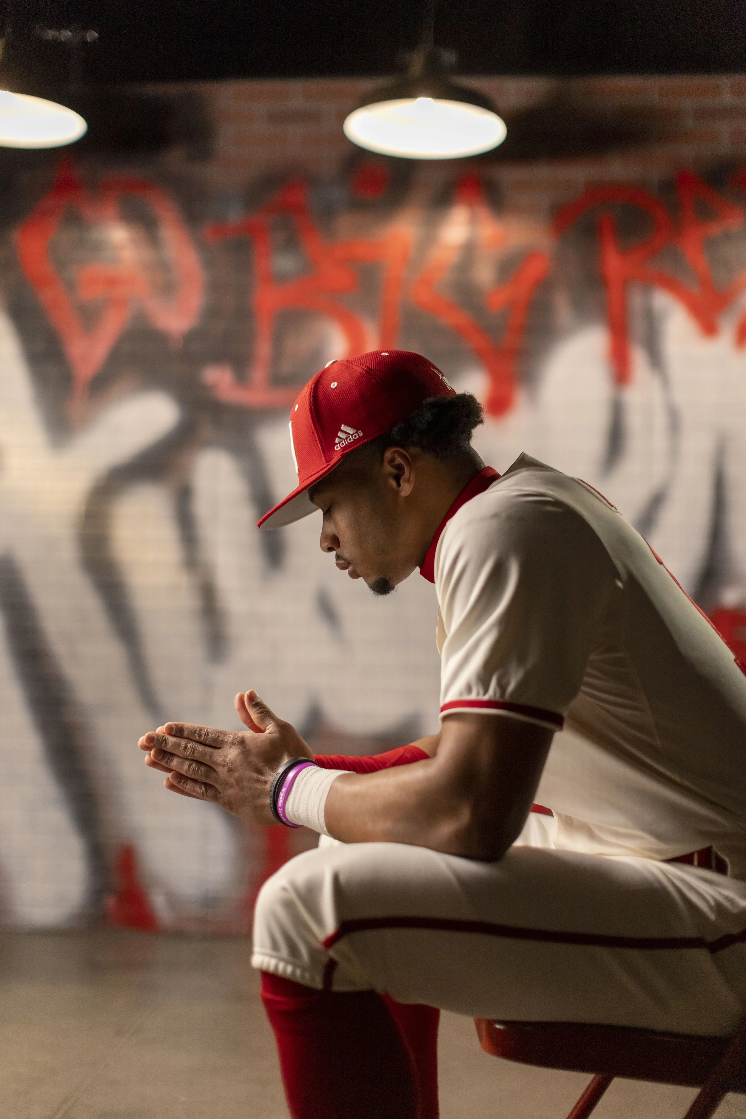 A baseball player in a white and red uniform sitting on a chair with his hands clasped together, wearing a red cap and a wristband, in front of a graffiti-covered brick wall.