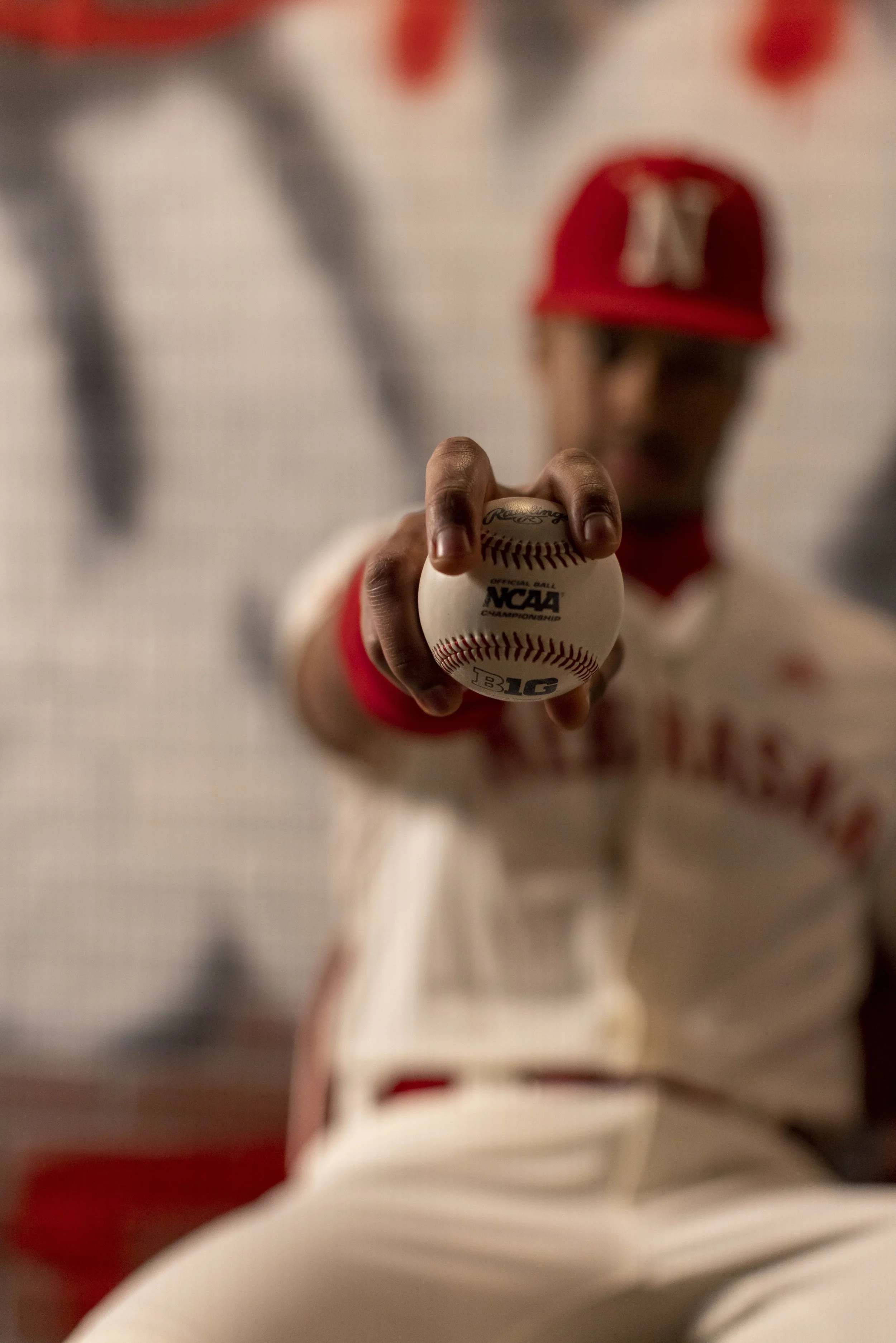 A young baseball player in uniform and a red cap holding a baseball towards the camera, with a blurred background.