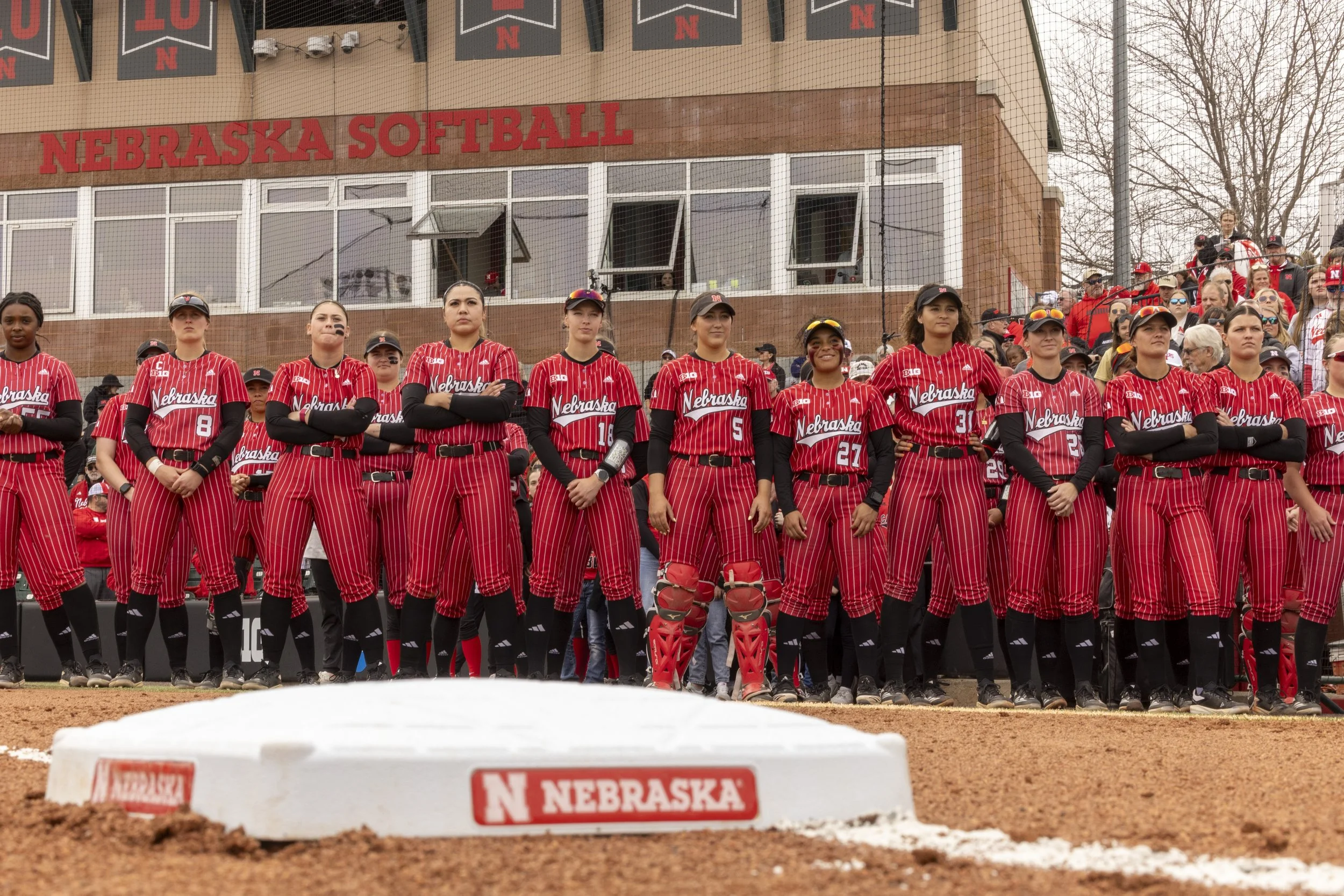Female softball players standing in a row on the field during a game, wearing red and black striped uniforms with 'Nebraska' written across their jerseys. They are at the edge of the field near the pitcher's mound, with spectators in the background a