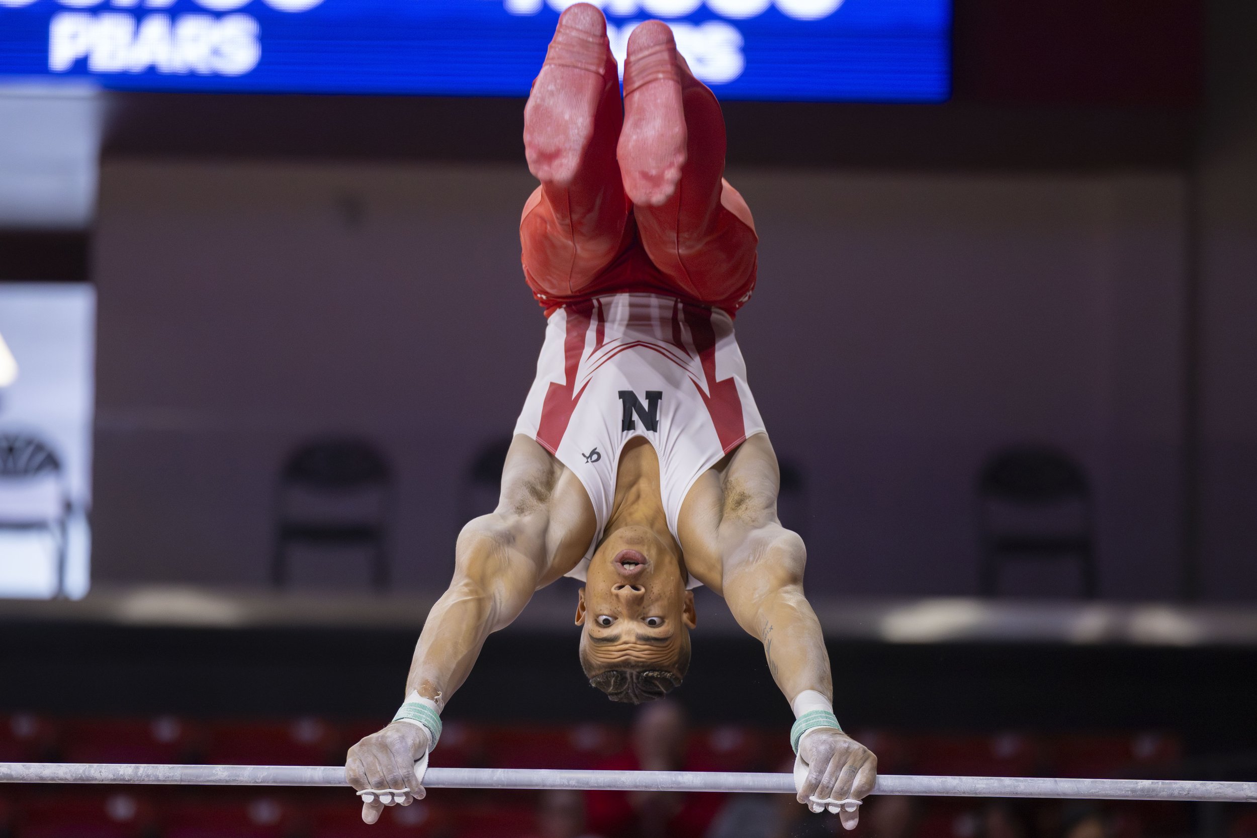 An athlete performing a handstand on a gymnastics balance beam, wearing a white and red uniform with the letter 'N' on it.