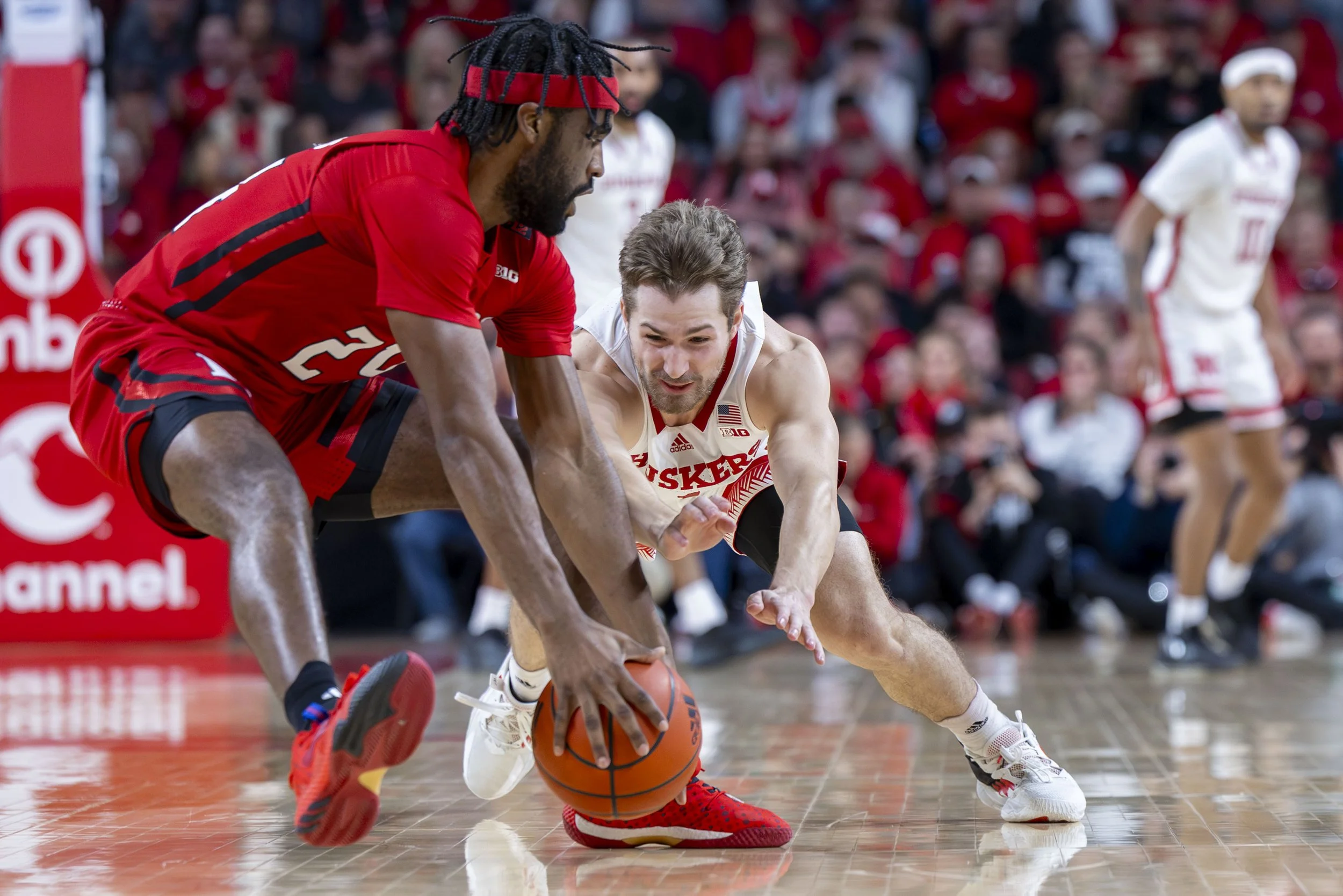Two basketball players reaching for a loose ball on the court during a game, with an audience in the background.