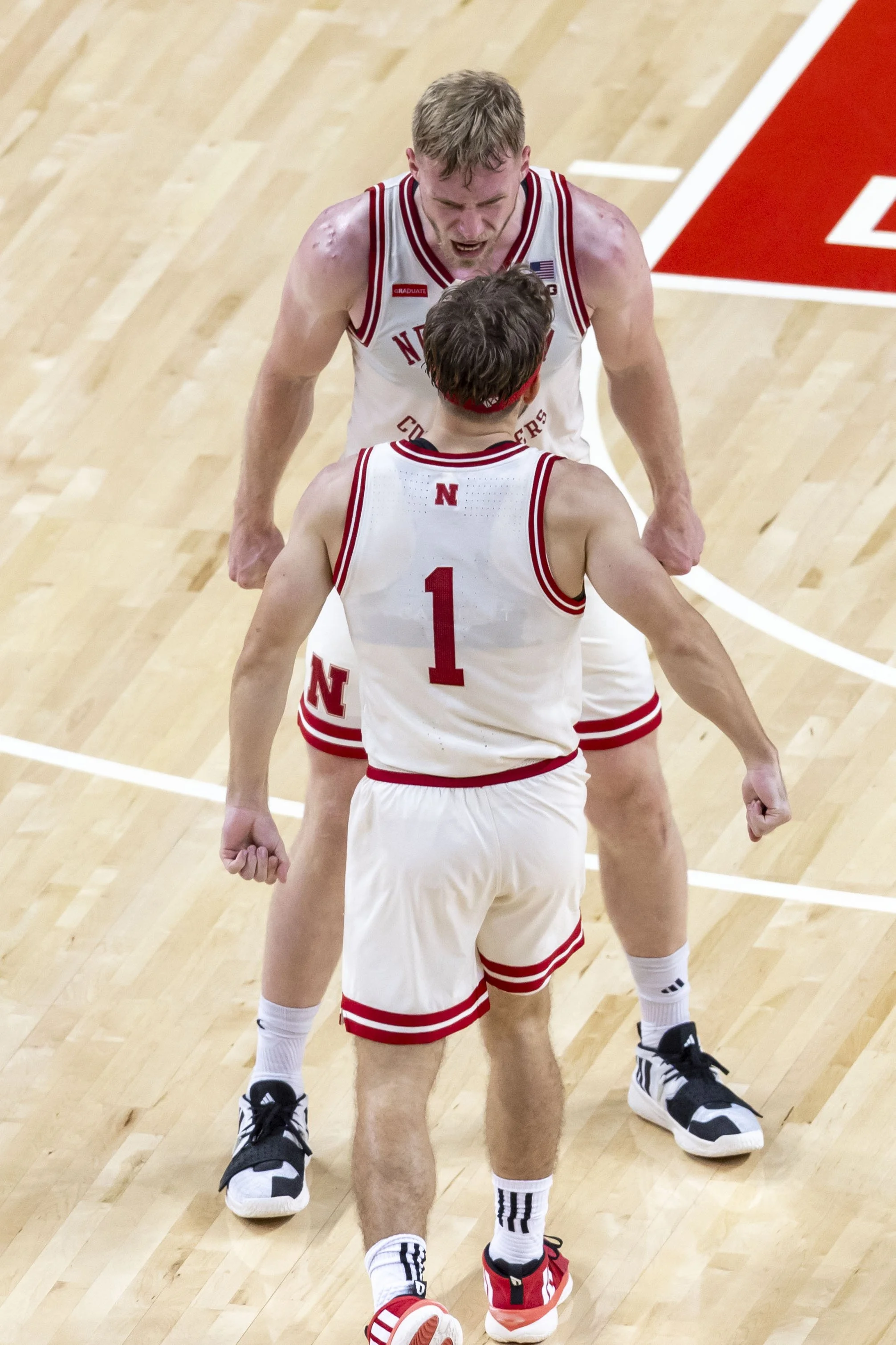 Two basketball players in white uniforms with red accents are celebrating on a wooden court, with one player looking angry and the other with his back to the camera.