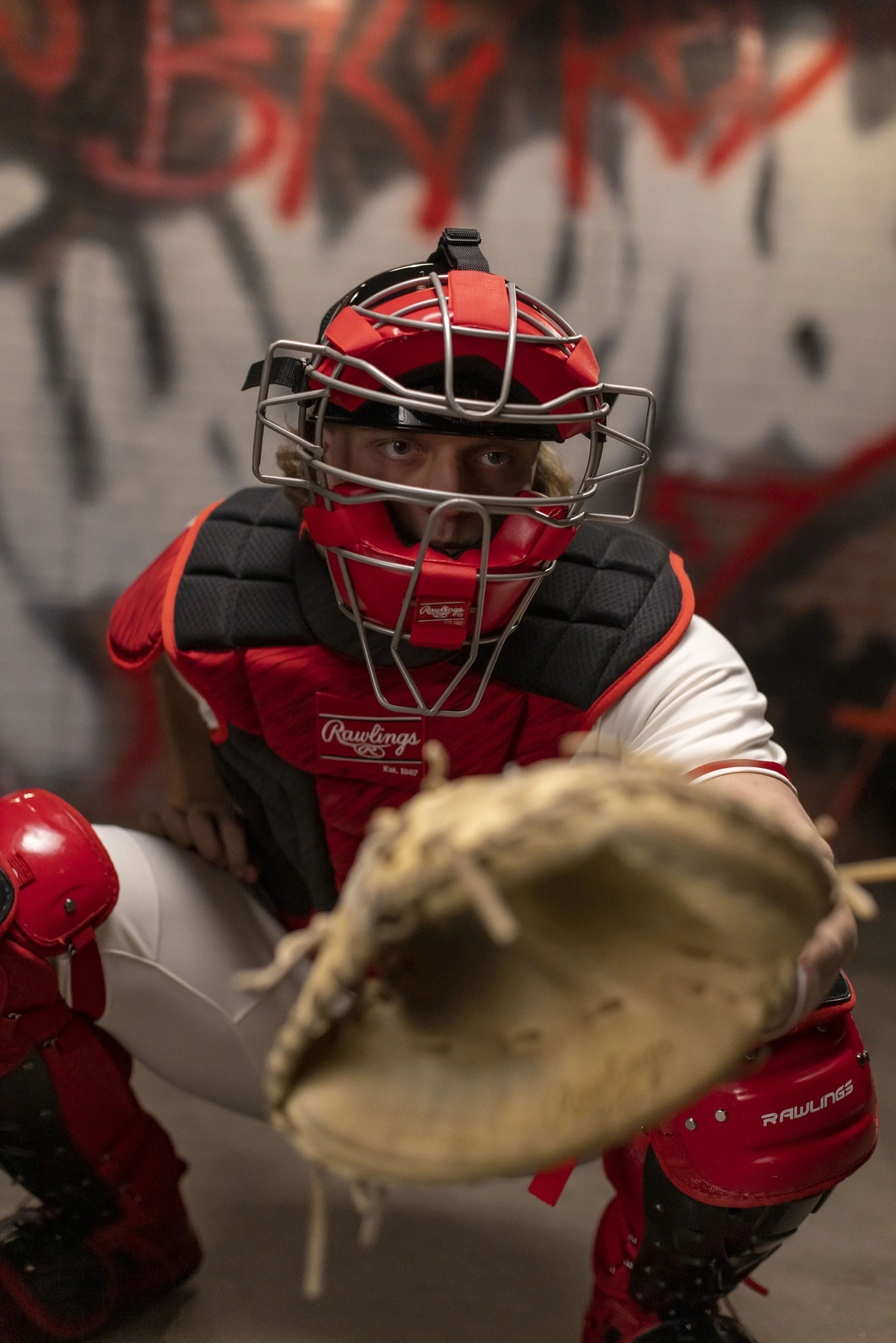 A baseball catcher in full gear crouching with a glove extended forward, set against a graffiti-covered wall.