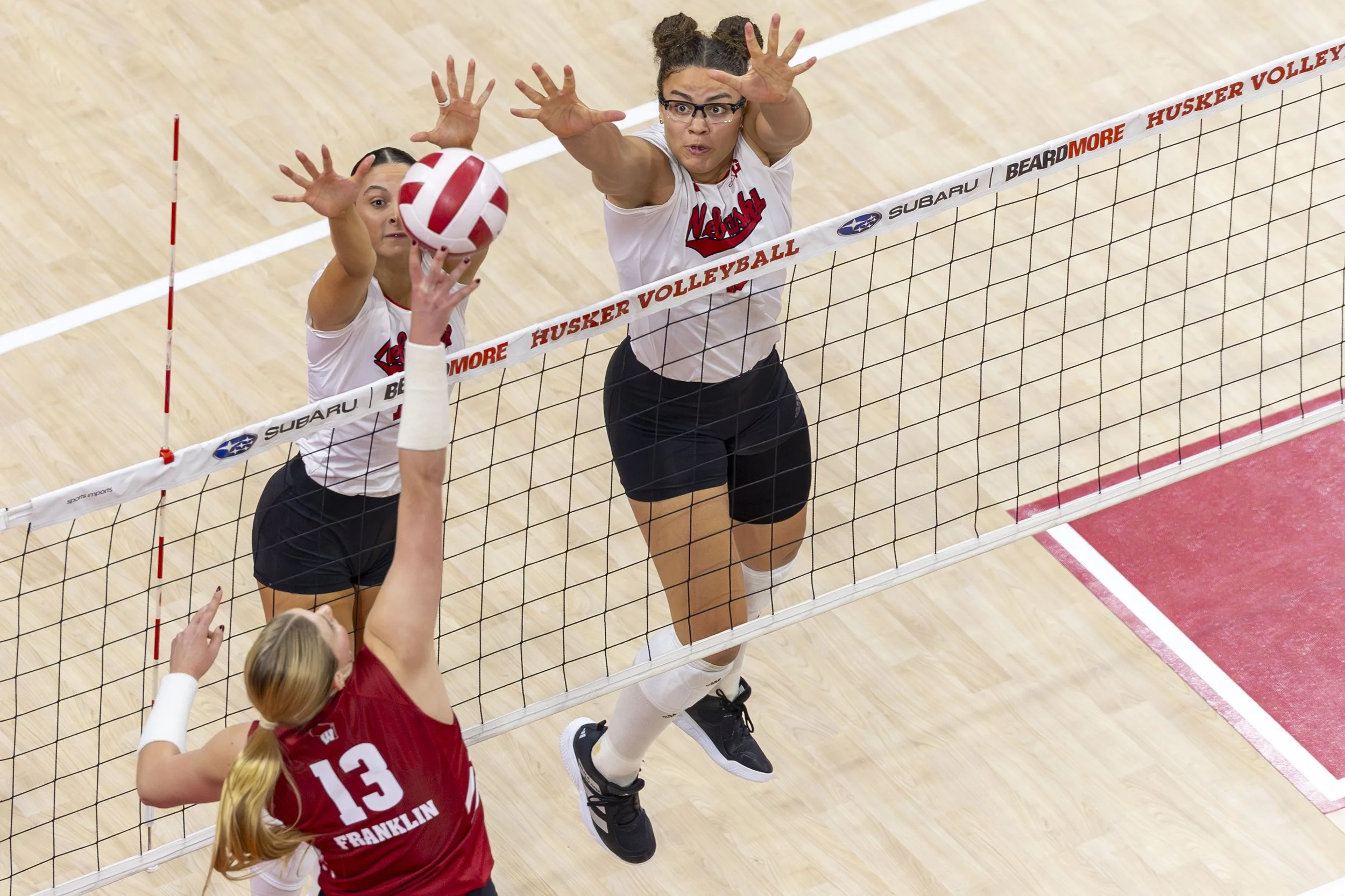 Two volleyball players from Nebraska Huskers and Franklin teams jump to block and spike the ball during a match in a volleyball court.