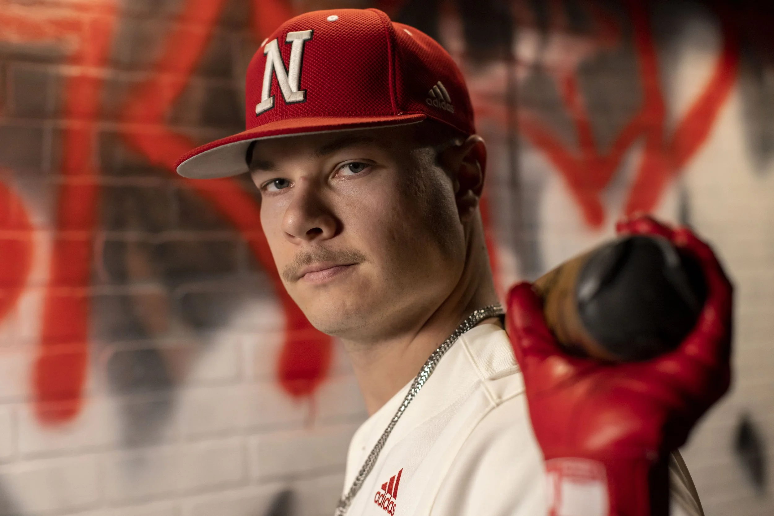 A young man wearing a red baseball cap with an 'N' on it, a white Adidas t-shirt, and a silver chain, holding a baseball bat over his shoulder with a red glove, standing in front of a brick wall with red graffiti.